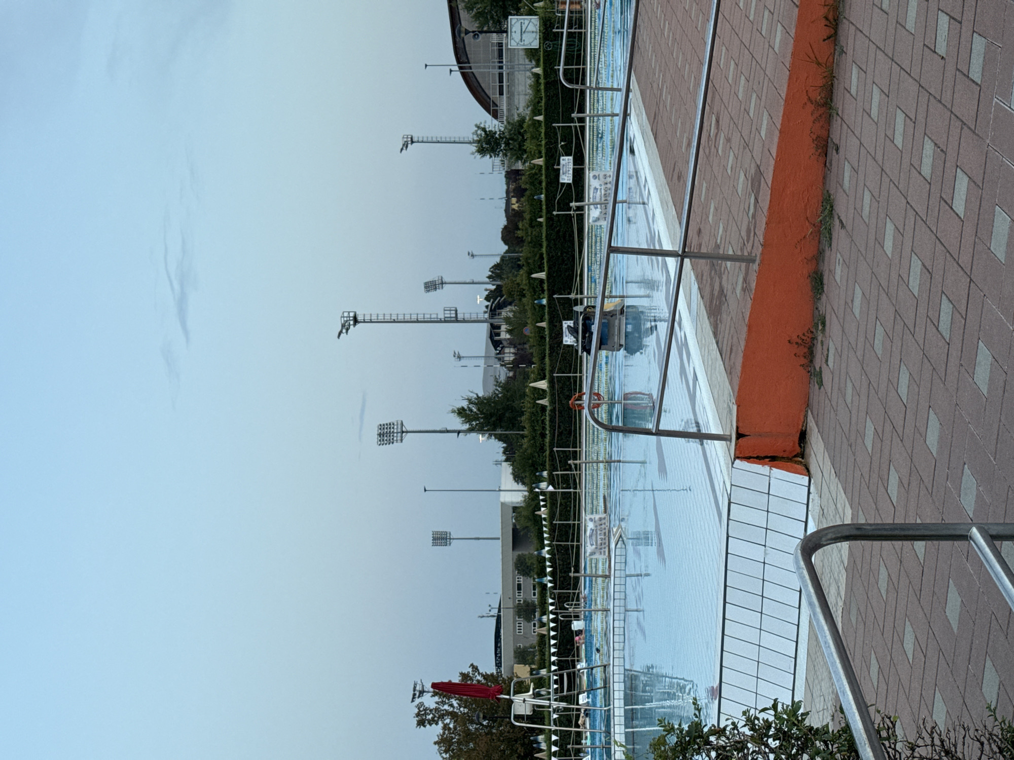 The image depicts an outdoor swimming pool facility in Parma, Italy, likely at dusk, given the soft lighting and clear sky. The main subject is a large, rectangular swimming pool filled with clear blue water. The pool has marked lanes visible on its bottom and is surrounded by a paved area made of reddish-brown bricks laid in a diagonal pattern.
To the left of the pool, a lifeguard chair with a red umbrella is positioned, suggesting it is a public facility. Several people are visible in the pool, appearing to be swimming or relaxing. A red life preserver ring is also visible near the edge of the pool.
In the background, several tall light poles with floodlights are erected, indicating the facility can be used after dark. Behind the pool area, there is dense green vegetation and some buildings, including what appears to be a sports complex with a curved roof. The overall environment suggests a recreational area within a city.