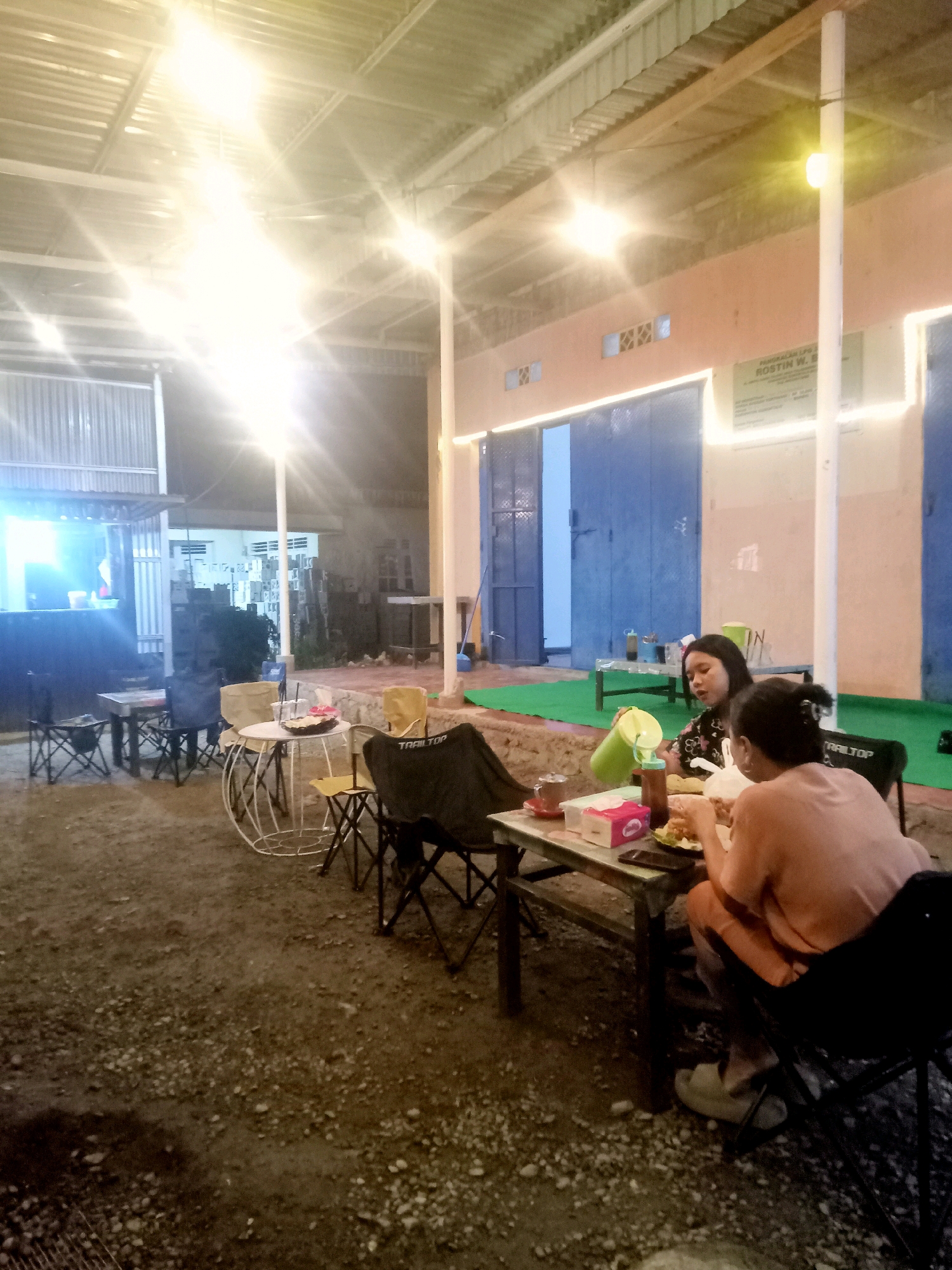This image captures an outdoor seating area at night, likely a casual eatery or cafe, possibly located in the city of Mongolato, Indonesia. String lights illuminate the scene, casting a warm glow on the corrugated metal roof overhead and the surrounding environment. The ground is covered with gravel and dirt, with several tables and foldable chairs scattered about.
Two women are seated at a table in the foreground, engaged in eating. One woman, in the foreground and facing away from the camera, wears a light pinkish-orange shirt and dark pants, with light-colored slippers on her feet. She is actively eating, with food on her plate and a fork in her hand. The second woman, to her left and facing more towards the camera, has dark hair and is wearing a dark floral-patterned top. She is holding a green pitcher and appears to be pouring a drink. A pink tissue box and a bottle of sauce are visible on their table, along with plates of food and cups.
In the background, there are more tables and chairs, a building with large blue doors, and what appears to be a small food stall or kiosk with bright lights. A sign is visible on the building wall, bearing text in Indonesian. The overall atmosphere suggests a relaxed evening setting for dining and socializing.