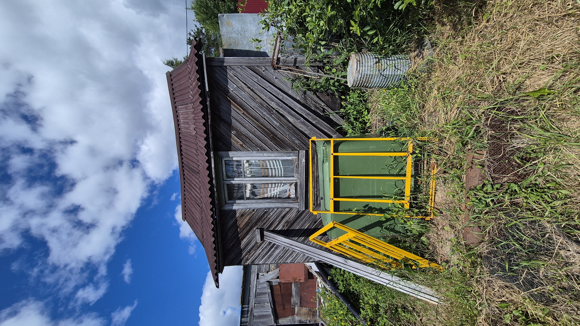 The image depicts an exterior scene in the rural settlement of Mashezero, Russia. The central focus is a weathered, dark wooden shed with a corrugated metal roof. A single window with striped curtains is visible on the shed's facade. In front of the shed, a large green trash bin is partially enclosed by a bright yellow metal frame. To the left, a section of a wooden structure, possibly another shed or fence, is leaning against the main shed. Various elements of disuse and overgrowth are present, including tall grass, weeds, a rusted metal barrel, and scattered debris. The sky above is bright blue with scattered white clouds. The overall impression is one of a neglected rural property on a clear day.