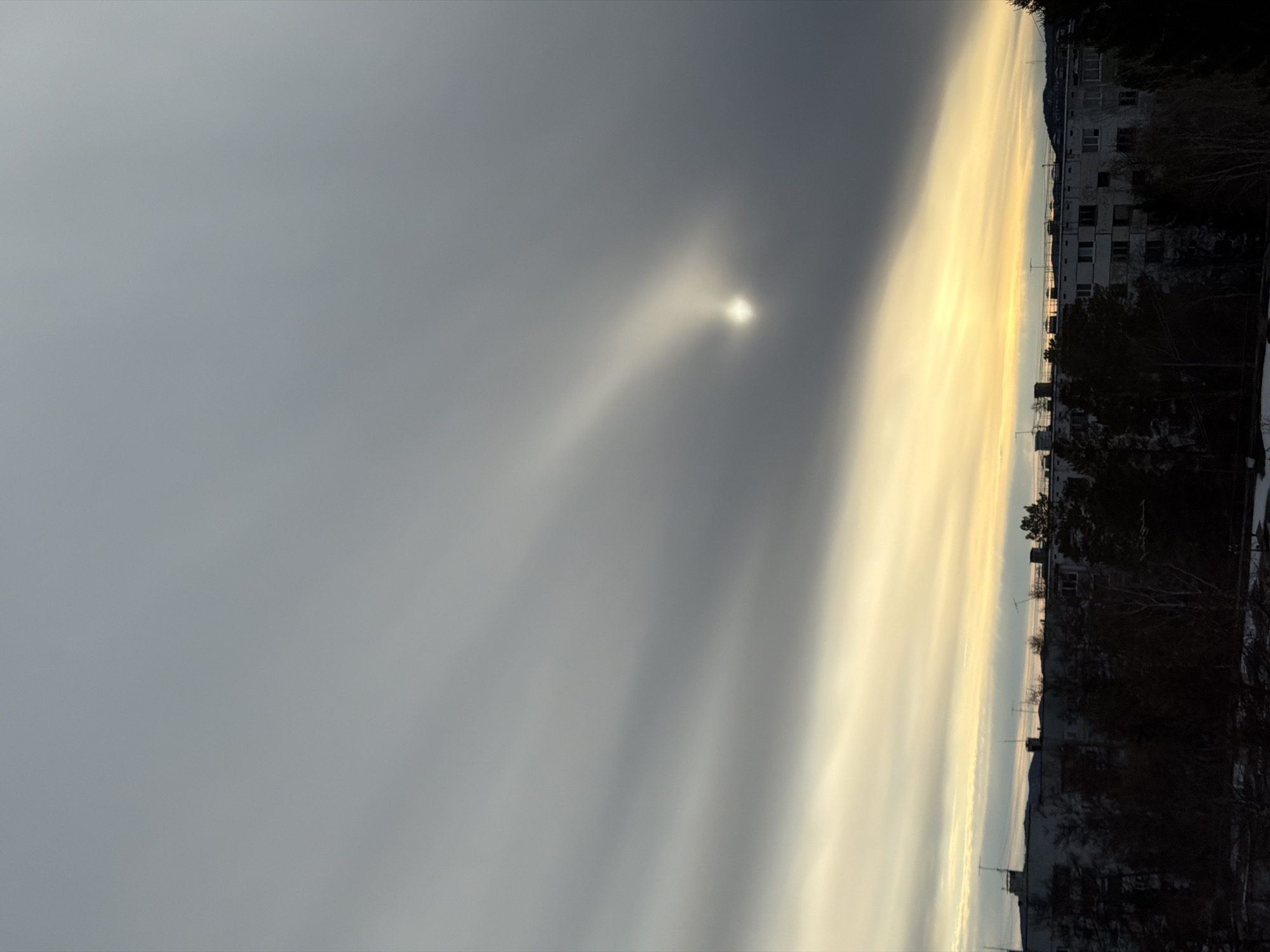 The image shows a view in Ust-Ilimsk, Russia, at dusk or dawn. A large, multi-story building is visible on the right side of the frame, partly obscured by trees, some of which are bare. The sky is mostly overcast with varying shades of gray, but there is a significant break in the clouds where the sun appears, creating bright, yellowish-orange streaks that illuminate the horizon. The sun's glare creates a halo effect in the sky. The ground is not clearly visible.