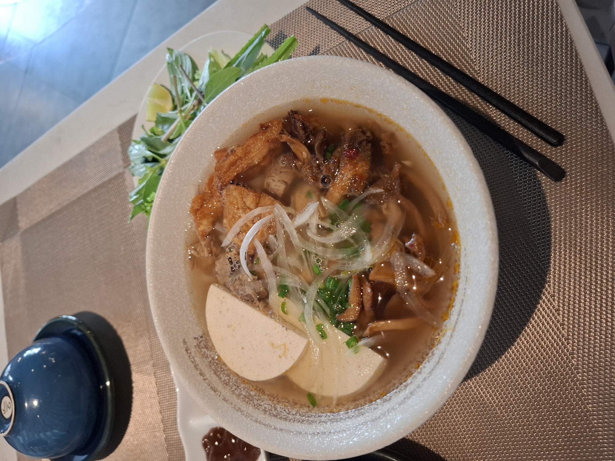 A top-down view shows a bowl of pho in Nha Trang, Vietnam. The pho is served in a white bowl and contains noodles, meat, and what appears to be a slice of Vietnamese ham. A small dish of dipping sauce is to the left of the bowl, and a plate of herbs and lime is to the right. Two black chopsticks rest on the table beside the bowl. In the background, a blue lid sits on a small dish, and part of a white table and a textured placemat are visible. The lighting suggests an indoor setting, possibly a restaurant.