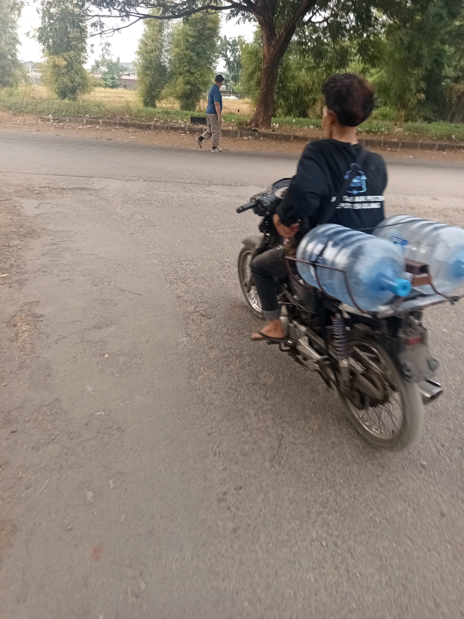 In an outdoor urban setting in West Jakarta, Indonesia, a person is riding a motorcycle. The motorcycle is equipped to carry two large, blue-capped water jugs, which are secured to the rear of the vehicle. The rider is wearing a black long-sleeved shirt and dark pants, and their feet are visible in flip-flops. In the background, another person walks along the side of a road. The environment includes a paved road, grassy areas, and trees. The lighting suggests it is daytime.