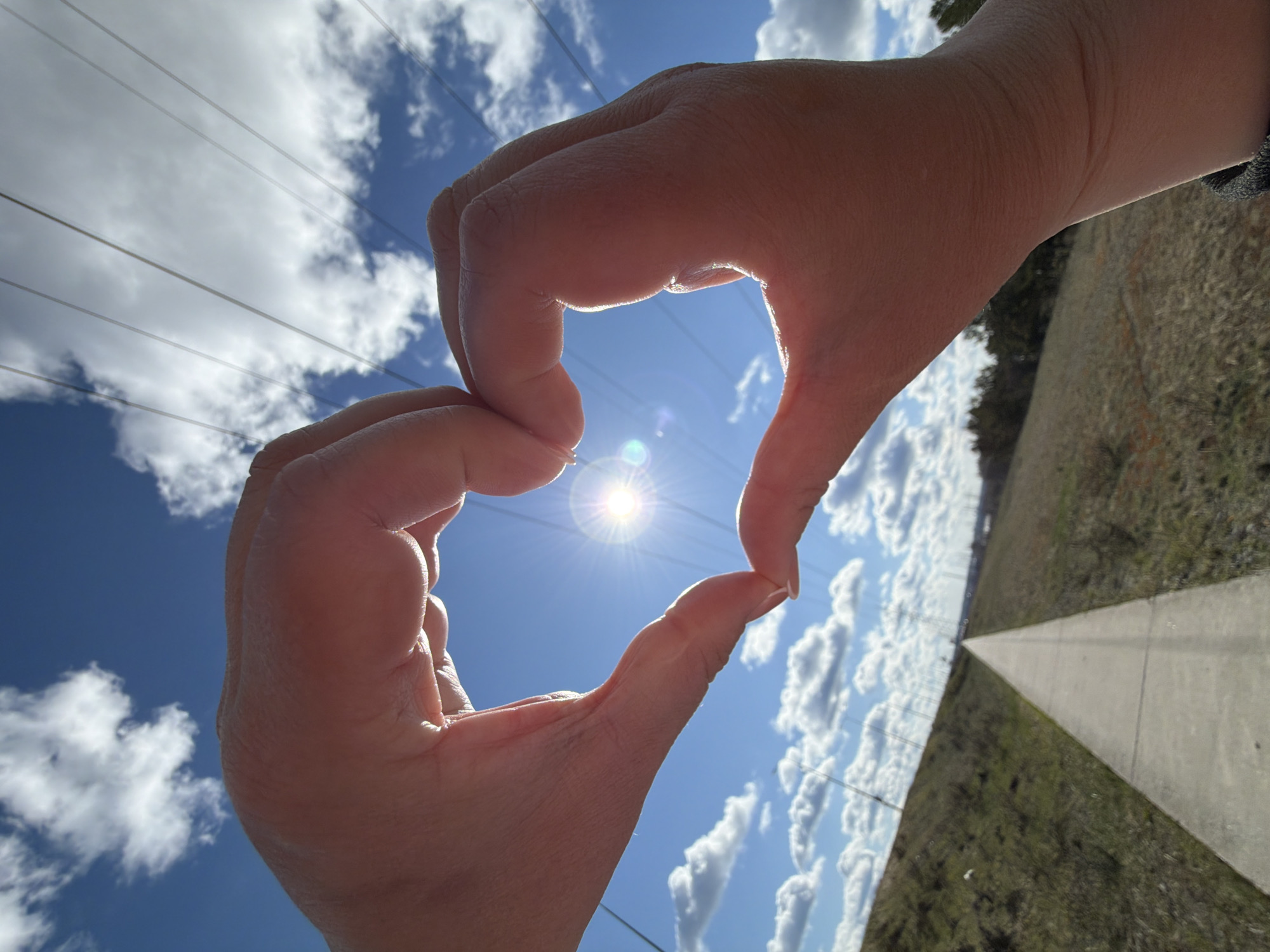 The image presents an outdoor scene in Chaikovsky, Russia, characterized by bright daylight and a clear, blue sky adorned with scattered white cumulus clouds. The central focus is a person's two hands, positioned to form a distinct heart shape against the sky. Within the heart's opening, the sun shines brightly, exhibiting a prominent starburst effect, making it the radiant focal point of the composition.
Several thin power lines or utility cables are visible stretching horizontally across the upper and left portions of the sky. Below the horizon line, especially towards the right, the landscape consists of dry, earthy grass or vegetation. A straight concrete path or road recedes into the distance on the right side of the frame, hinting at a pathway through the terrain.
The photograph is taken from a low angle, looking upwards towards the sky and the sun, creating an expansive feel. The strong sunlight illuminates the hands, emphasizing their form and texture. The image has a slightly tilted perspective, adding to its dynamic quality. There is no visible text. The overall mood conveyed by the heart gesture and the sunny weather is one of warmth, love, and optimism.