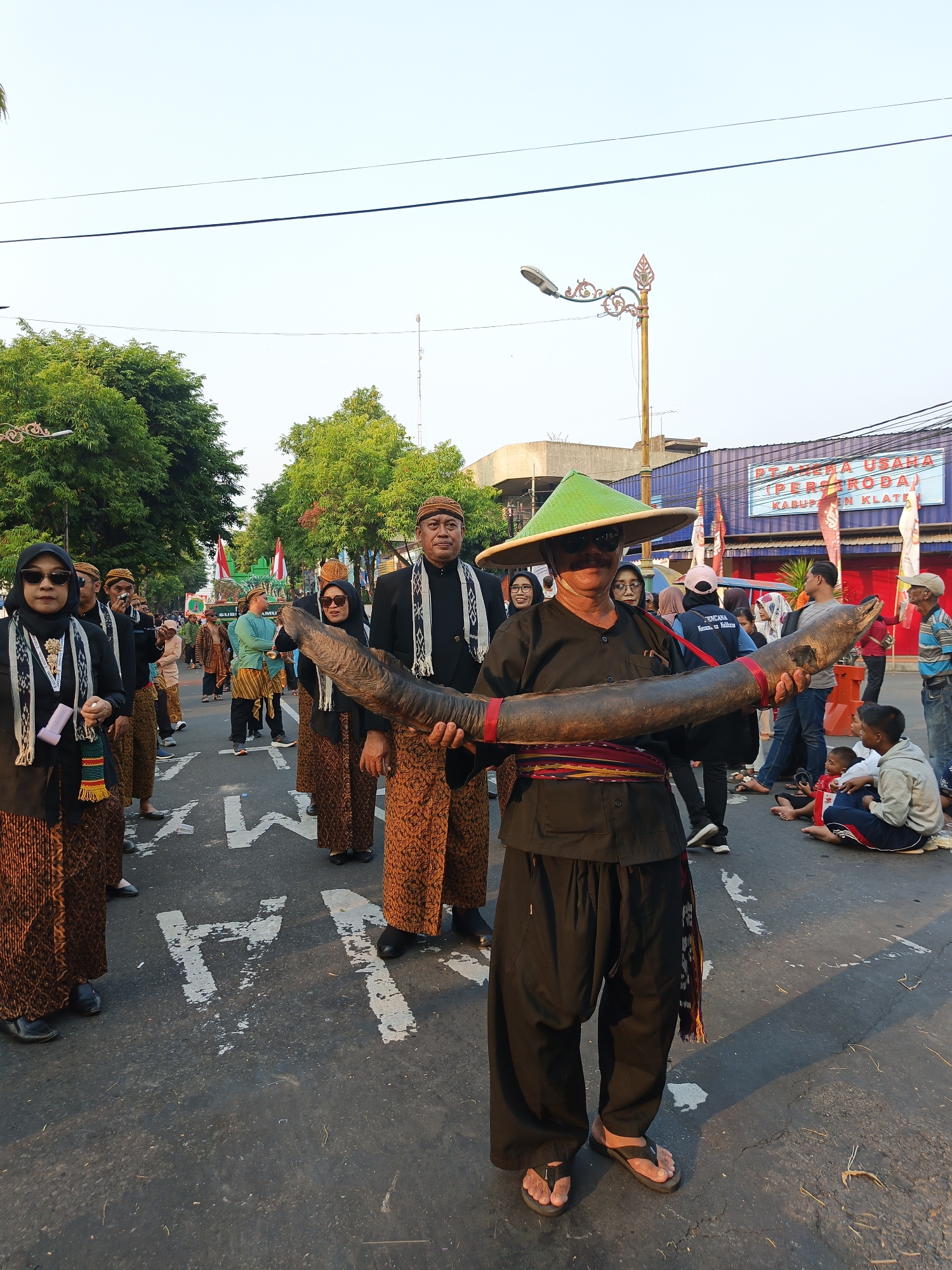 In Klaten, Indonesia, a man in traditional attire, including a green conical hat and a black outfit with loose pants and sandals, proudly holds a large, dark, horn-like object. He is positioned centrally in the frame, facing the camera. He is surrounded by other individuals also dressed in traditional Indonesian clothing, many wearing batik skirts and headscarves. The scene appears to be part of a procession or cultural event, indicated by the presence of multiple people walking in an organized manner on a paved road. Some spectators, including children, are seated on the side of the road, observing the event. In the background, buildings and trees line the street, and a banner with text that includes "PT. ANGGRA USAHA PERKASA (PERKASA) KABUPATEN KLATEN" is visible, further confirming the location. The overall atmosphere suggests a lively community celebration.
