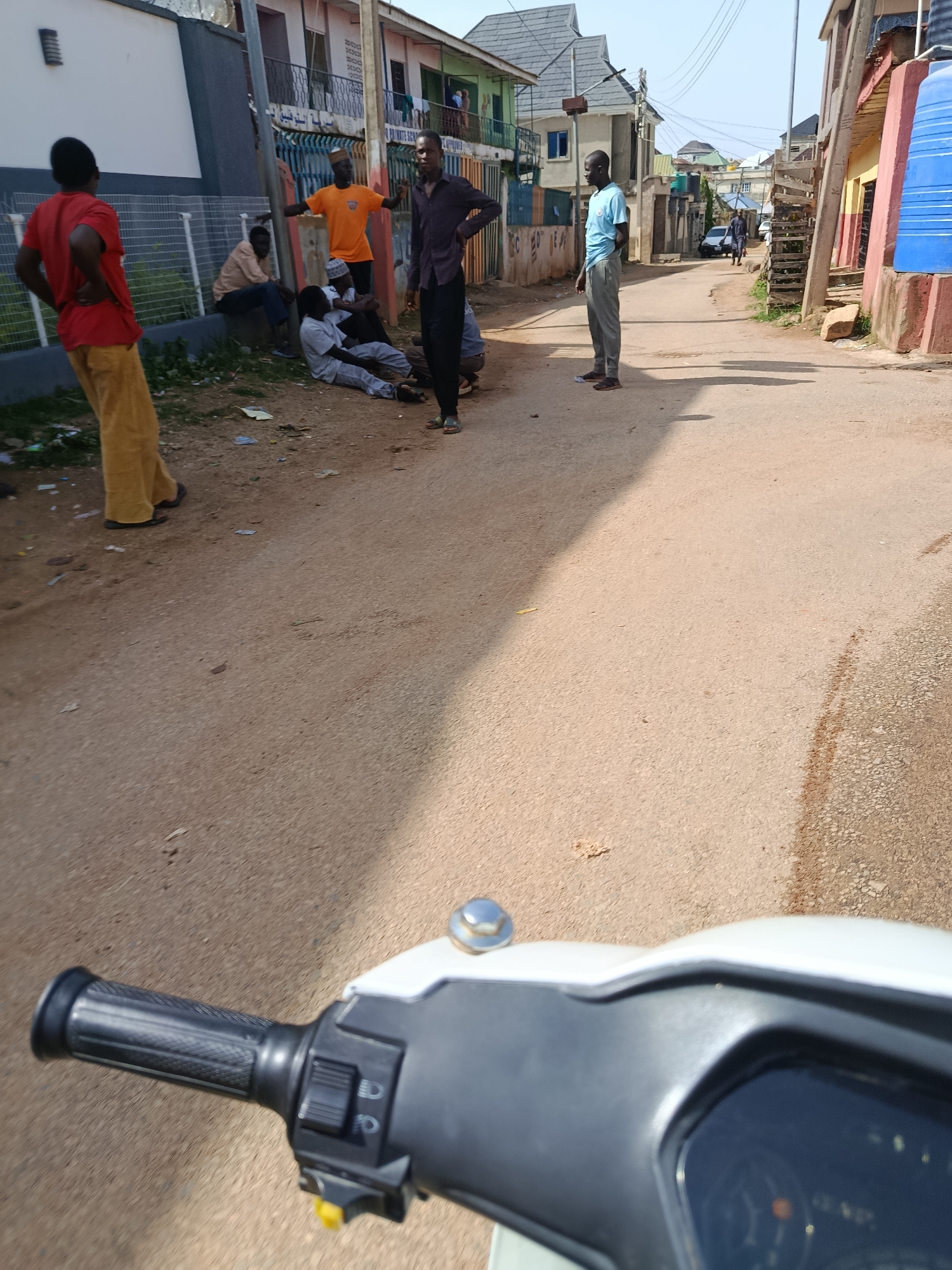 The image depicts a street scene in Rafin Pa, Nigeria, with several individuals gathered. In the foreground, a motorcycle's handlebars and part of its dashboard are visible, suggesting the photo was taken from the perspective of someone riding it. On the left side of the street, a person in a red shirt and yellow pants stands facing away from the camera. Nearby, several other individuals are seated or standing, appearing to converse or observe. One man in an orange shirt is standing with his arms outstretched, while another in a dark shirt and black pants stands with his hands on his hips. Further down the street, two men in light blue shirts and grey pants are visible, one standing and looking towards the camera, and another walking away. The street is paved with asphalt and lined with buildings, some of which appear to be residential with multiple stories. Power lines are strung overhead. The overall impression is a snapshot of everyday life in a Nigerian urban setting.