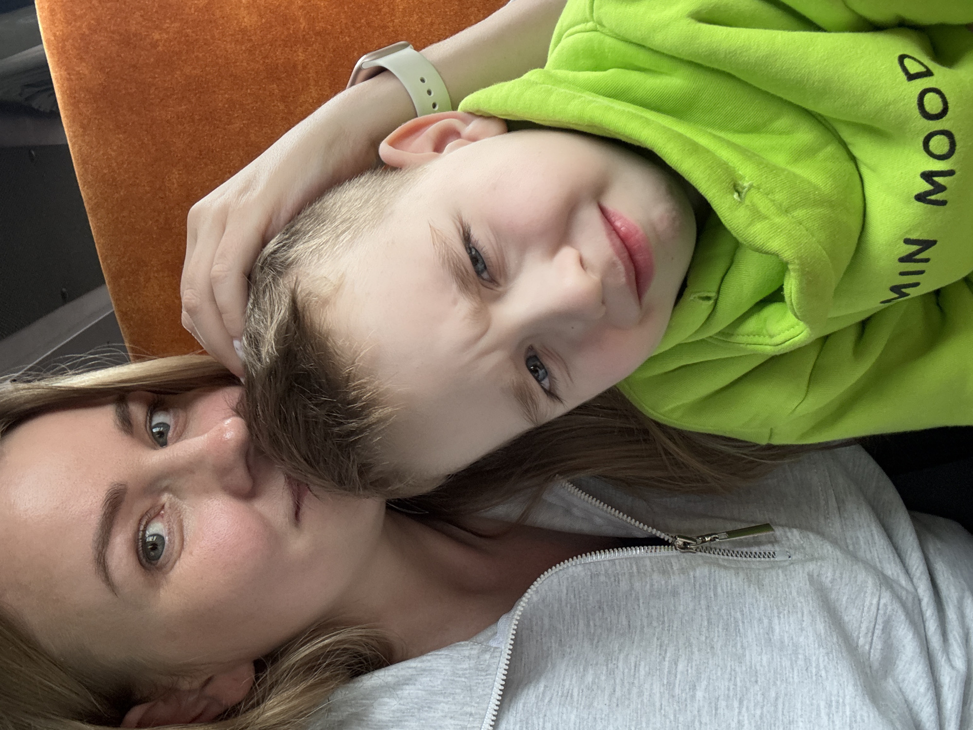 A woman and a young boy are captured in a close-up indoor portrait, potentially within the city of Chaikovsky, Russia. The woman, positioned on the left, has blonde hair, fair skin, and light eyes. She wears a light grey half-zip hooded sweatshirt and exhibits a gentle expression while looking towards the viewer. Her left hand is placed on the head of a young boy in the foreground. The boy, with light hair, presents a distressed facial expression, characterized by a frown, downturned mouth, and visibly sad eyes, directly facing the camera. He is wearing a bright lime green hooded sweatshirt with partial black text on the front, reading "MIN MOOD," and a light-colored smartwatch on his left wrist. The background consists of a textured orange surface, suggestive of upholstery, and a dark, indistinct upper section, indicative of an indoor setting.