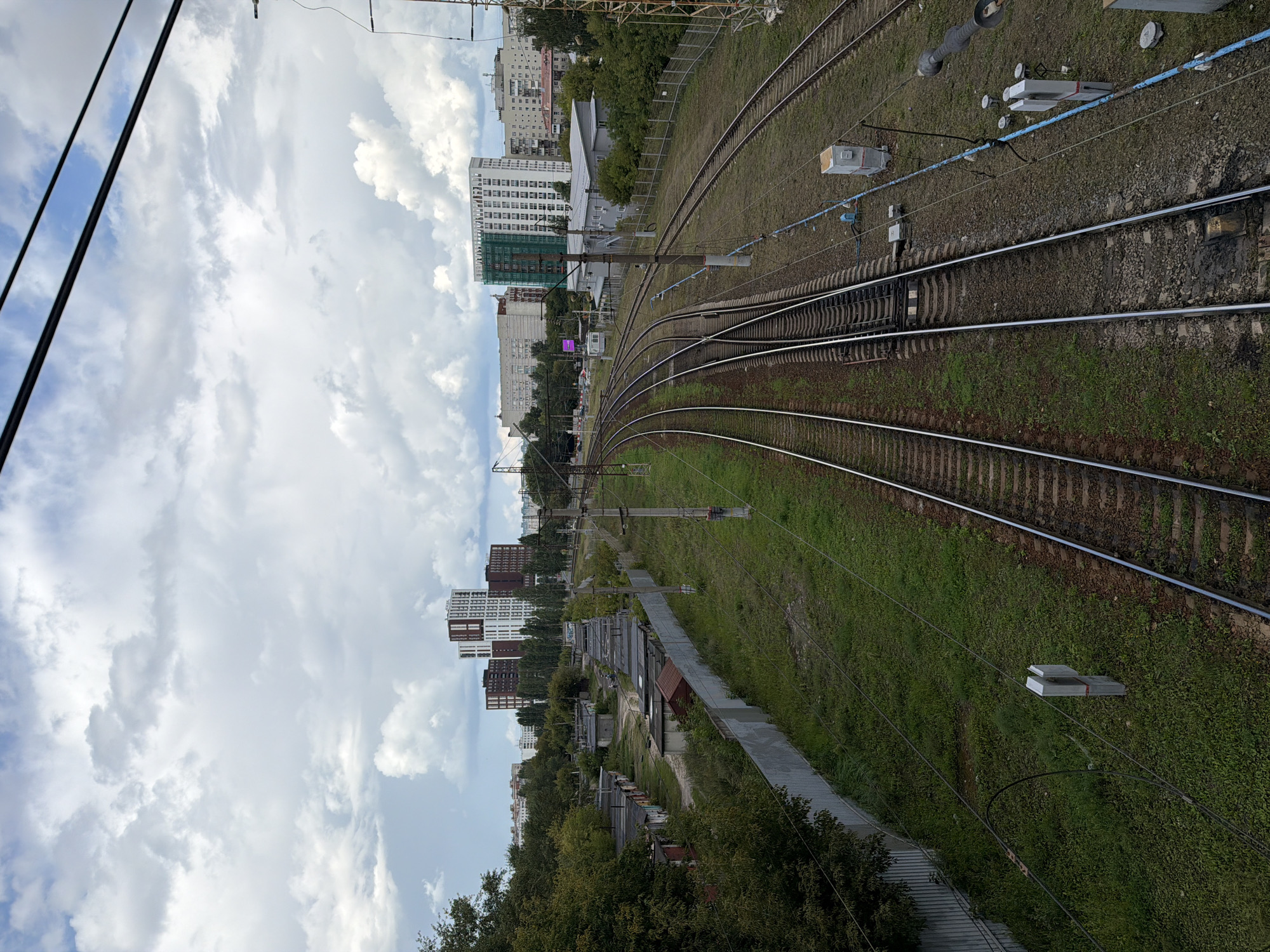 The image shows a view of multiple train tracks with overhead electrical wires stretching into the distance. In the foreground, the tracks are laid on a gravel bed with grassy areas on either side. A grey metal fence runs along the left side of the tracks, separating them from a row of small, weathered garages. Beyond the garages, a line of trees is visible, and further in the distance, several modern multi-story buildings rise above the tree line, including two prominent towers with a mix of brick and white facades, and another tall white building partially covered in green scaffolding. The sky is cloudy with patches of blue visible. The scene appears to be an industrial or semi-industrial area within a city, likely on the outskirts, given the combination of railway infrastructure, older garages, and newer residential or commercial buildings. This location is identified as being near the railway station in the Kanavino district of Nizhny Novgorod, Russia, specifically in the vicinity of the former "Gorodskoy Rynok" (City Market) area which has undergone significant redevelopment with new construction. There are no people clearly visible in the image.