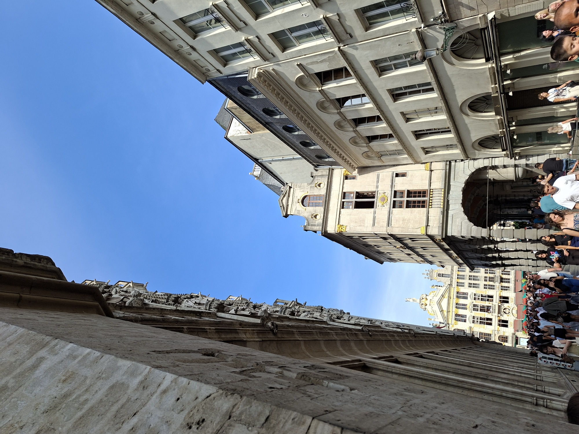 This is a street-level, slightly upward-angled photograph taken in Brussels, Belgium, likely on the Grand Place. The foreground is dominated by the textured stone facade of a building on the left, casting a strong shadow and obscuring much of the left side of the image. The sky is a clear, vibrant blue, indicating fair weather.
In the mid-ground, a street is bustling with pedestrians walking in various directions. The architecture of the surrounding buildings is varied but exhibits a European urban style, with light-colored stone or stucco facades, numerous windows, and ornate architectural details. The building in the far background, with its bright white and gold embellishments, is a prominent landmark, likely the Town Hall or one of the guildhalls on the Grand Place.
To the right of the foreground building, a series of buildings line the street. These structures have multiple stories, with windows of varying shapes and sizes, some arched and some rectangular. Decorative elements like cornices, moldings, and small balconies are visible. A black streetlamp with a green decorative arm is mounted on one of these buildings.
The people in the image are diverse, dressed in casual, warm-weather clothing. They are engaged in activities typical of a public square or busy street: walking, talking, and looking around. The overall impression is one of a lively, historic urban environment on a sunny day.