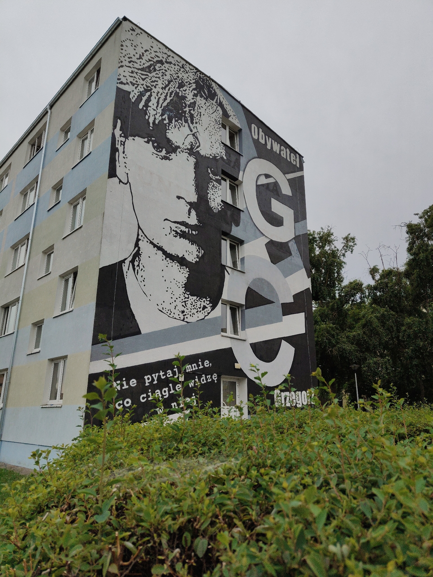 A mural covers the side of a multi-story building, depicting a large grayscale portrait of a man's face. The building is painted with horizontal bands of light blue, beige, and gray. To the right of the portrait, large white letters spell out "OBYWATEL" and "G C G", with "Grzego" partially visible at the bottom right. Below the portrait, in white text on a black background, are the words "nie pytaj mnie, co ciągle widzę w niej". The mural is partially obscured by lush green bushes in the foreground, with trees visible in the background to the right. The sky is overcast and gray. The location is Unknown, Unknown.