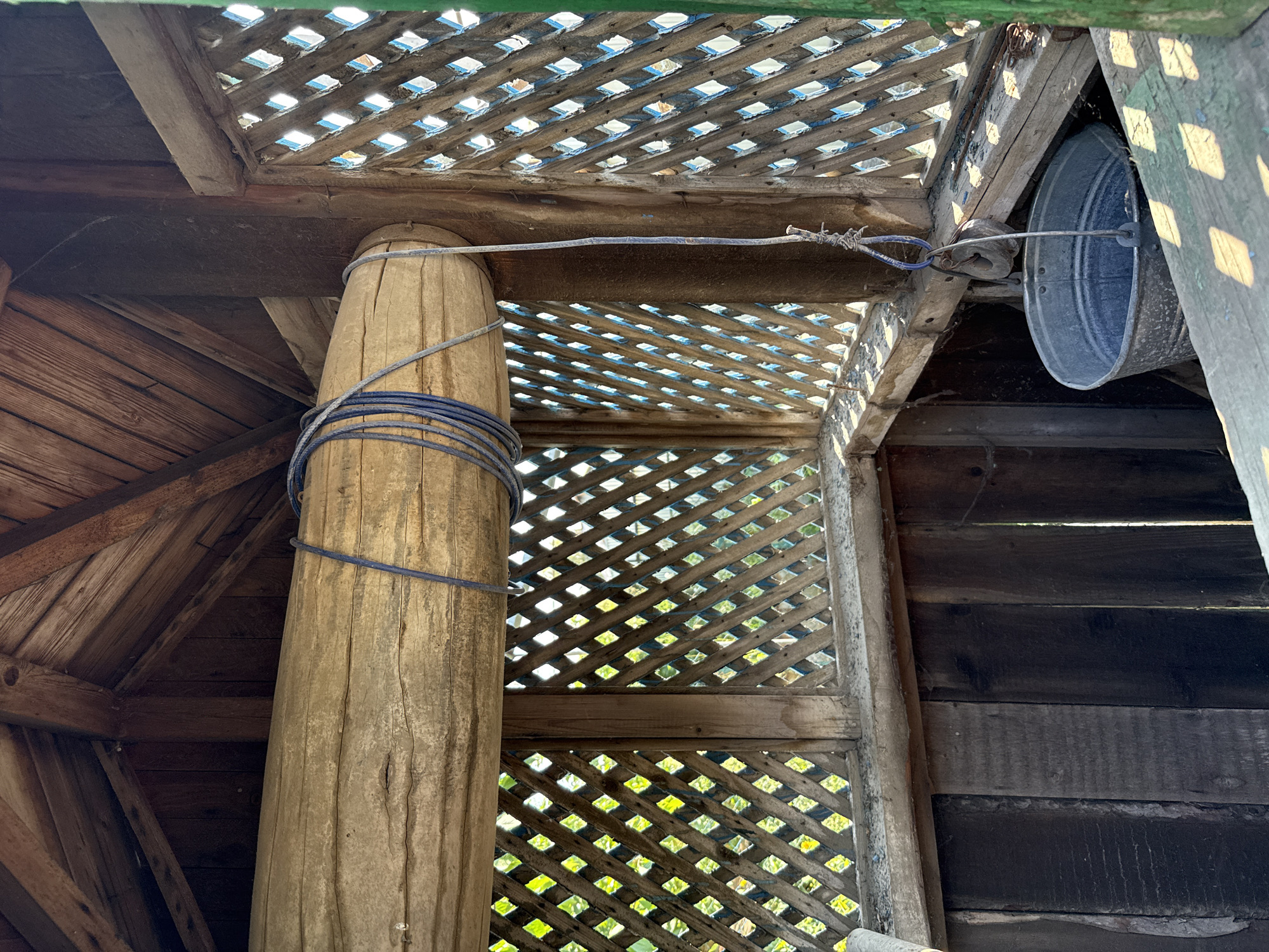 The image shows the interior of a wooden well structure. A large, rough-hewn wooden beam, likely a roller, is positioned horizontally. A blue rope is wrapped around this beam. A rope hangs down from the beam, attached to a metal pulley, which is in turn attached to a metal bucket. The bucket is partially visible at the bottom of the frame. The structure is made of rough-hewn wood, with lattice-like walls and a slatted roof. Sunlight filters through the lattice, casting dappled shadows. The environment appears rustic and outdoors, possibly in a rural setting. There are no people visible in the image.
