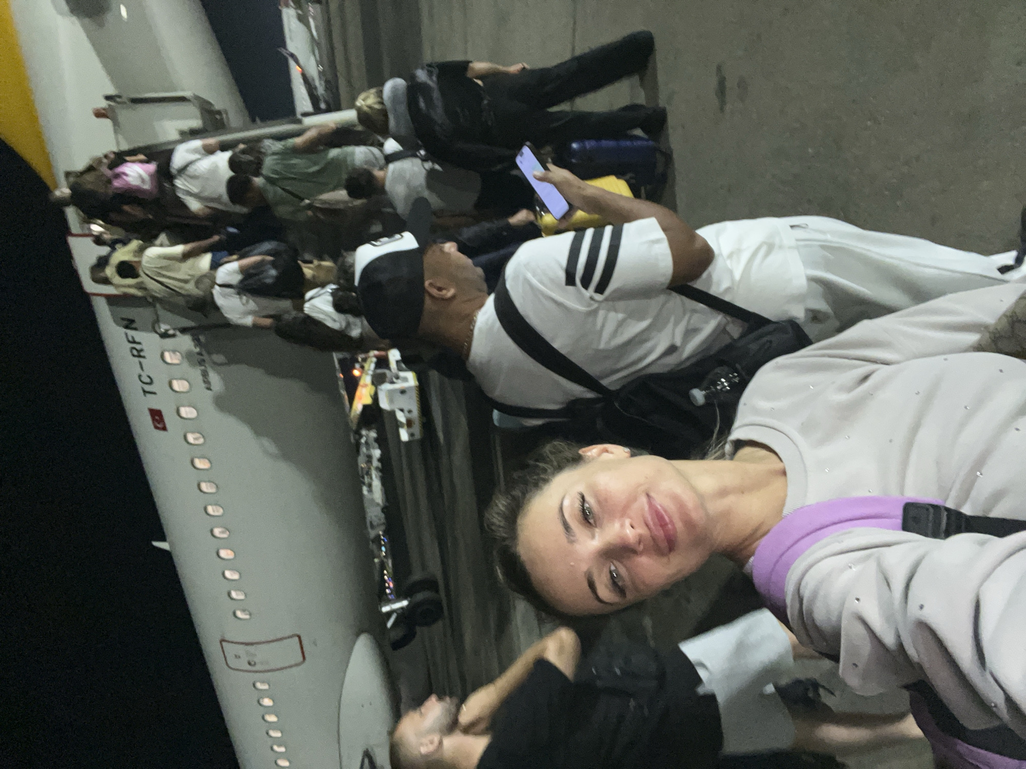 A group of people are boarding an airplane at night. The airplane, identified by the registration "TC-RFN," appears to be at an airport in Sharm El Sheikh, Egypt, as indicated by the context. The main subject in the foreground is a woman smiling at the camera, wearing a light-colored sweatshirt and a purple backpack. Behind her, several other individuals are ascending the stairs to the plane. The scene is illuminated by artificial lighting, casting shadows on the tarmac. Luggage is visible throughout the image, and airport service vehicles can be seen in the background. The overall impression is one of travel and departure under nighttime conditions.