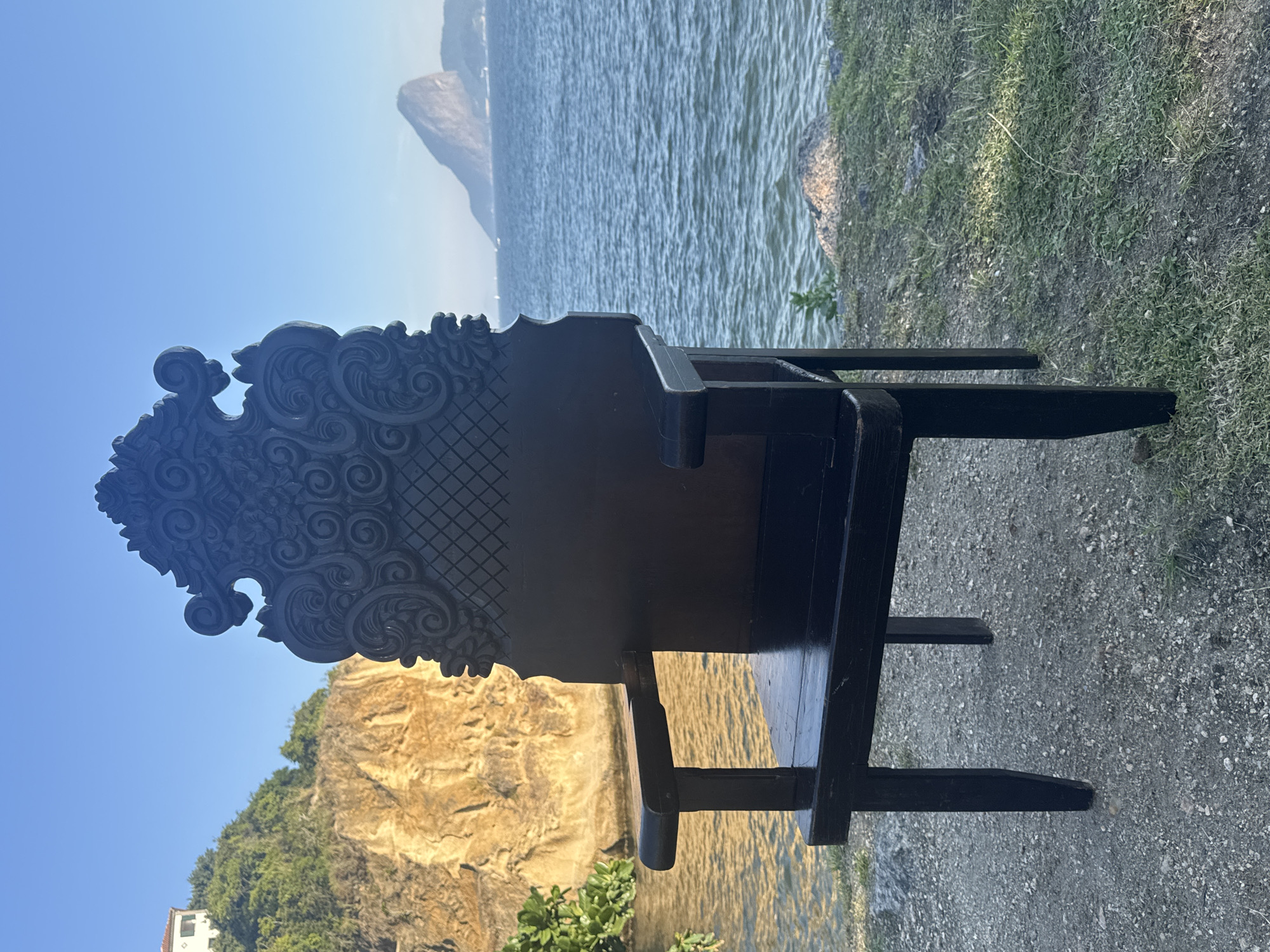An ornate, dark wooden chair sits on a gravel path next to a grassy area overlooking a body of water. In the background, a rugged, golden-hued cliff face rises from the water's edge, with lush green vegetation and a white building on its summit. Further in the distance, a distinctive, sugarloaf-shaped mountain rises from the sea under a clear blue sky. The scene is characteristic of the Boa Viagem neighborhood in Niterói, Brazil, offering a panoramic view of Guanabara Bay and the iconic Pão de Açúcar (Sugarloaf Mountain) in Rio de Janeiro.