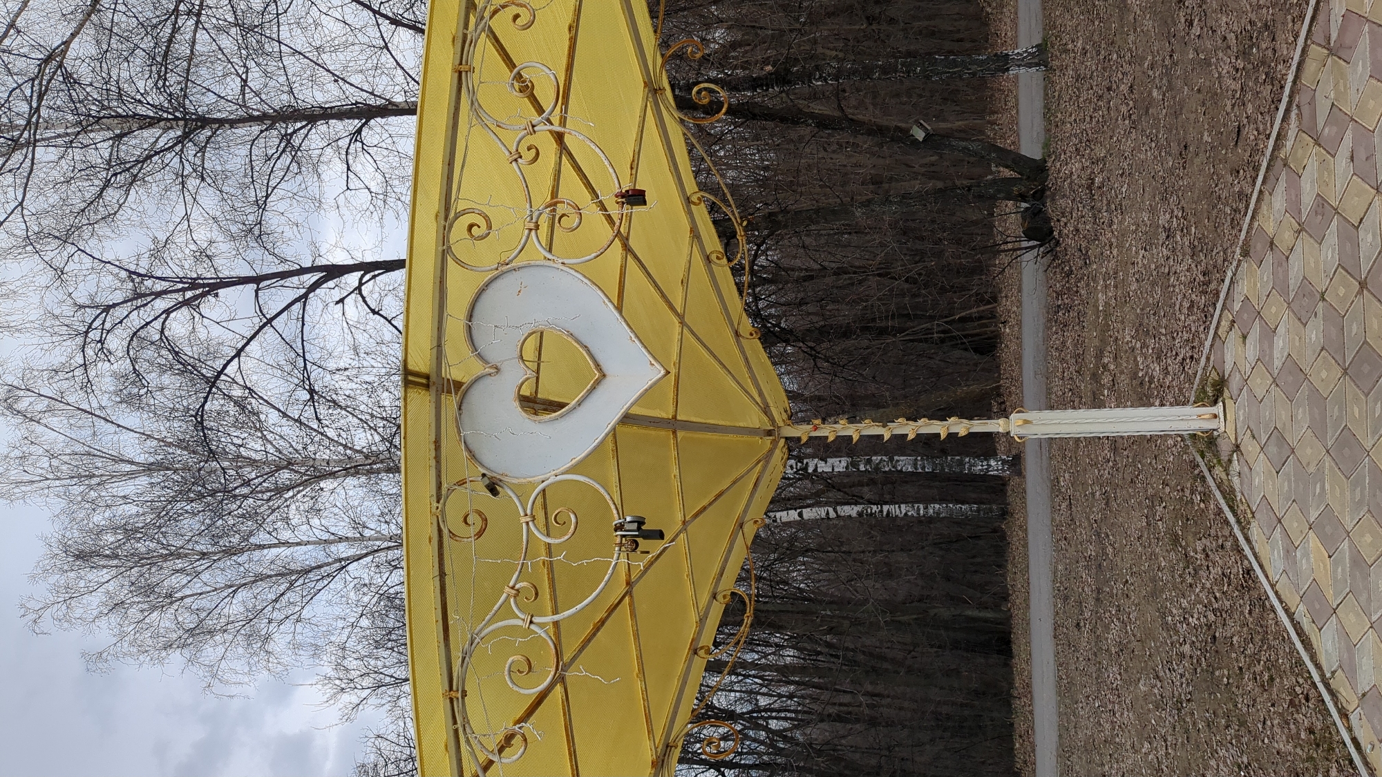 The image shows a decorative metal structure, likely an arch or gazebo, with a prominent white heart detail at its apex. The structure is painted yellow with ornate metalwork and positioned in a park-like setting. Bare trees suggest it is either late fall or early spring. A paved walkway runs alongside. The location is speculated to be Ulyanovsk, Russia. No people or actions are visible. The focus is on the architectural detail of the heart-shaped structure against the backdrop of trees and the surrounding landscape.