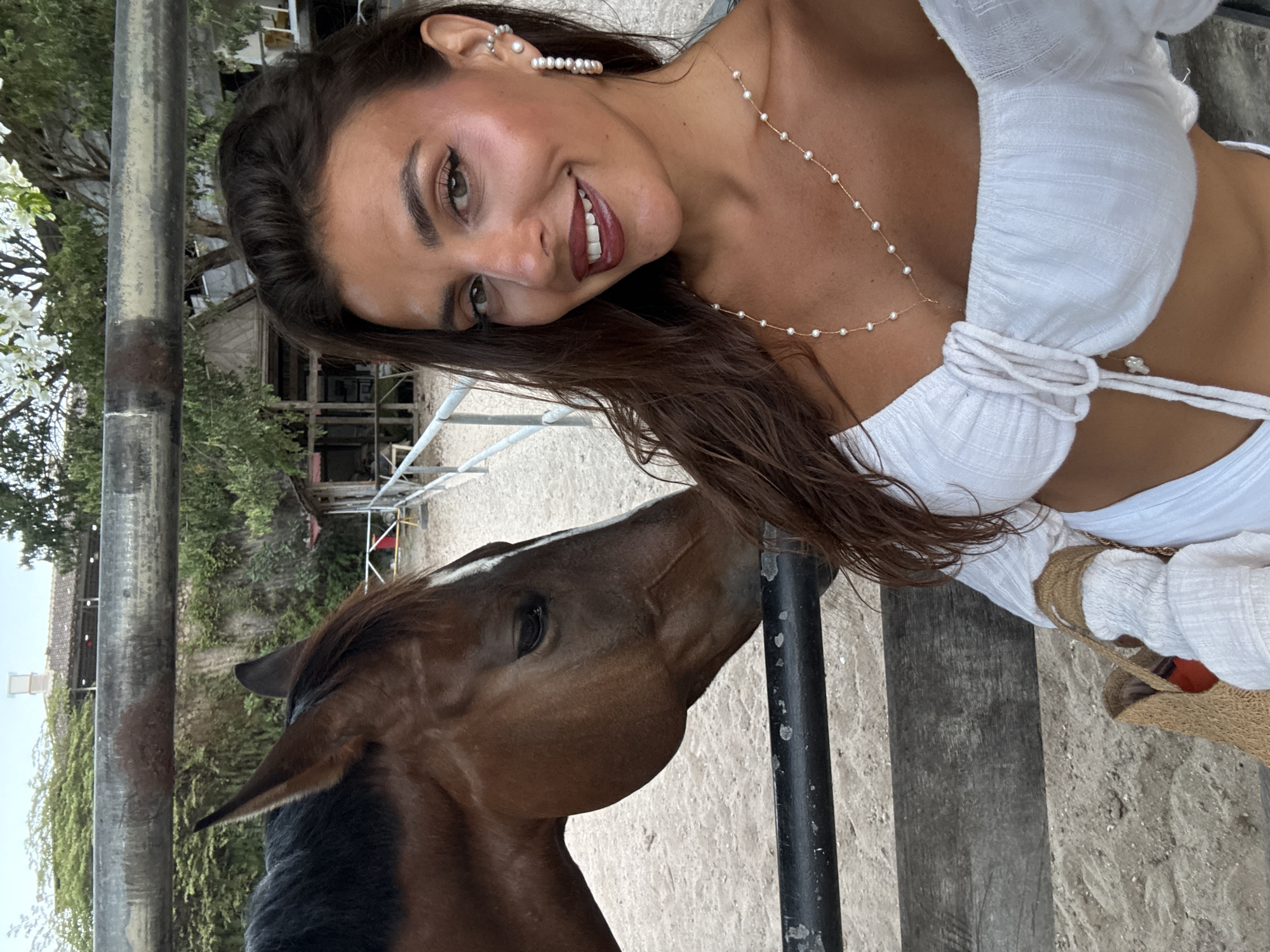 A woman is depicted in a close-up selfie alongside a brown horse in an outdoor, fenced enclosure in Kuta Selatan, Indonesia. The woman, positioned on the right, faces the camera with a smile, revealing upper teeth. She has long, dark brown hair, tanned skin, and wears a white off-the-shoulder crop top. Pearl jewelry includes a delicate gold necklace with small pearls, and multiple pearl earrings on her visible right ear (hoop, stud, and cuff). Her left arm is partially visible, holding a light brown woven bag. A dark metal fence runs horizontally, separating the woman from the horse. The horse, on the left, is brown with a white blaze on its face and its head is angled towards the woman, its eye appearing partially closed. The ground is sandy. In the background, additional metal and wooden fencing, dense green foliage, trees, and a distant light-colored building with a tiled roof and a central chimney-like structure are discernible under a light sky.