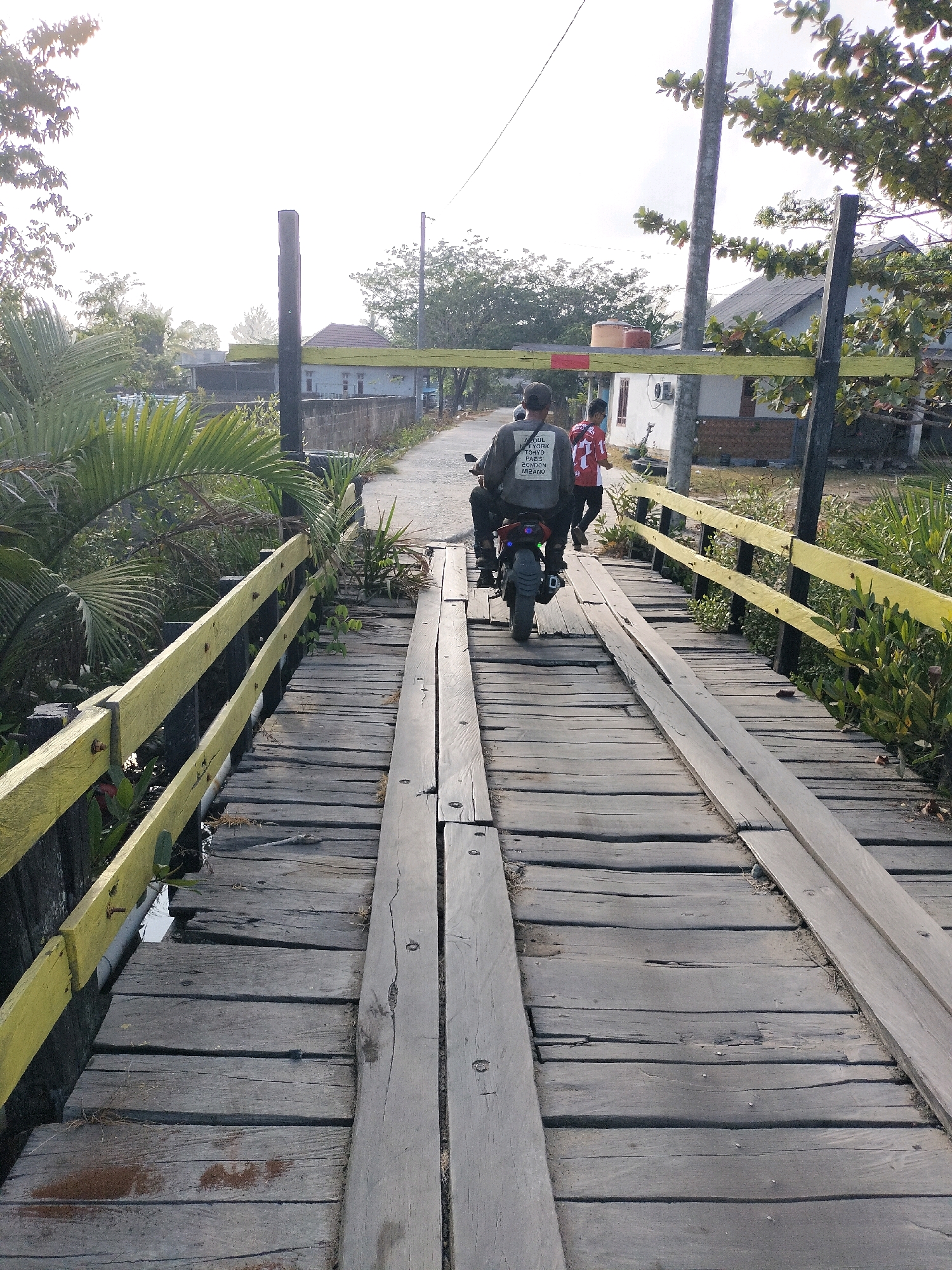 This image depicts a wooden bridge, likely in Bunati, Indonesia, with two individuals and a motorcycle. A person wearing a grey t-shirt with text that appears to be "SOUL NETWORK TOKYO PARIS LONDON MILANO" is riding a black motorcycle. Behind them, another individual in a red and white shirt is walking. The bridge has yellow-painted railings and shows signs of wear, with visible planks and some vegetation growing around its edges. The environment appears to be somewhat rural, with trees and buildings visible in the background. The sky is bright, suggesting daytime.