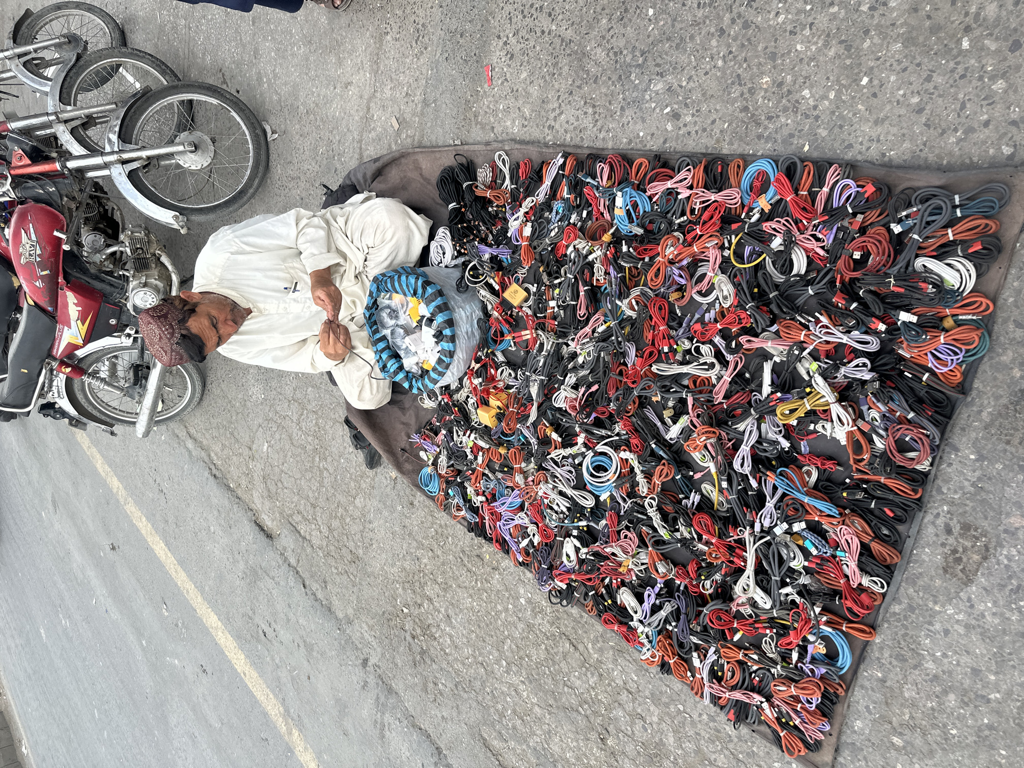 A man wearing a white shalwar kameez and a red patterned cap sits cross-legged on a dark brown cloth spread on an asphalt road. He is looking down and appears to be sorting through a small pile of black cables with his hands. In front of him, a large assortment of colorful cables, including red, black, white, blue, and pink, are spread out on the cloth, filling a significant portion of the frame. The cables are coiled and arranged in a seemingly disorganized manner. Behind the man, three motorcycles are parked side-by-side. The red motorcycle in the foreground has a license plate with the text "RAVI" visible. The scene appears to be a street market in Quetta City Tehsil, Pakistan, with the road surface showing signs of wear and tear. The overall lighting suggests it is daytime.