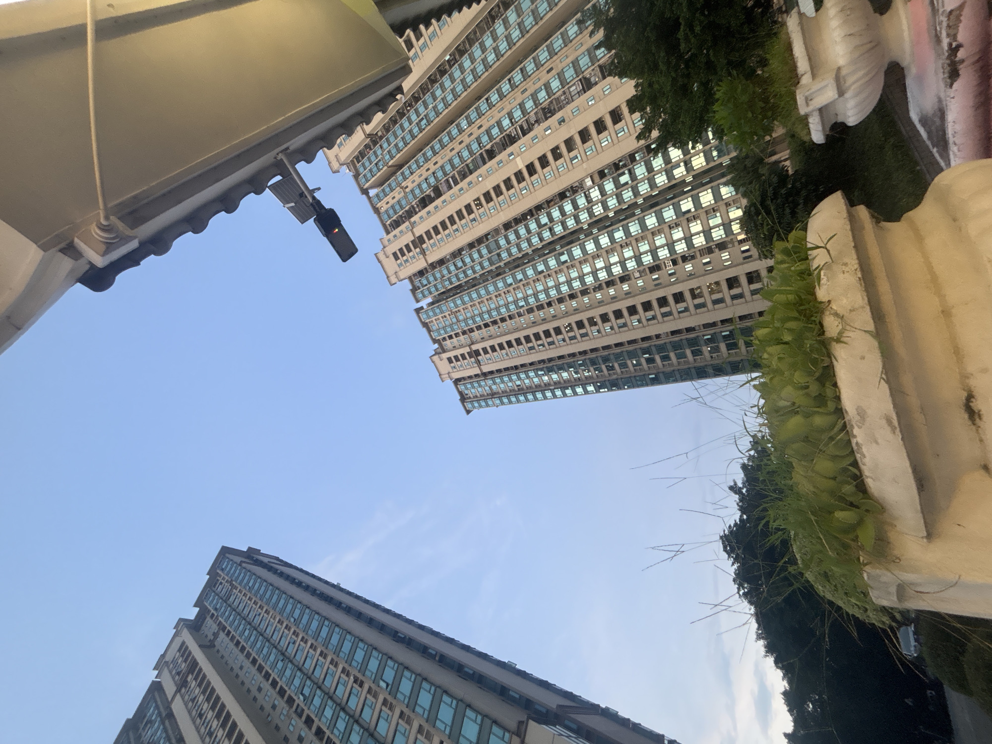 The image, captured in Nanhai District, China, presents an upward-angle view of two modern high-rise apartment buildings against a clear sky. The building on the left is taller and appears to be closer to the camera, its facade covered with rows of teal-tinted windows. The building on the right, slightly further away and to the right, has a similar design with numerous windows reflecting the light. In the foreground, a concrete planter with lush green plants and grass occupies the lower portion of the frame, partially obscuring the view of the buildings. A lamppost with a red light signal is mounted on an overhang in the upper right quadrant of the image, above the rightmost building. Dense foliage and trees are visible between the buildings and in the background, suggesting an urban setting with integrated greenery. The overall lighting indicates it may be dusk or early morning.