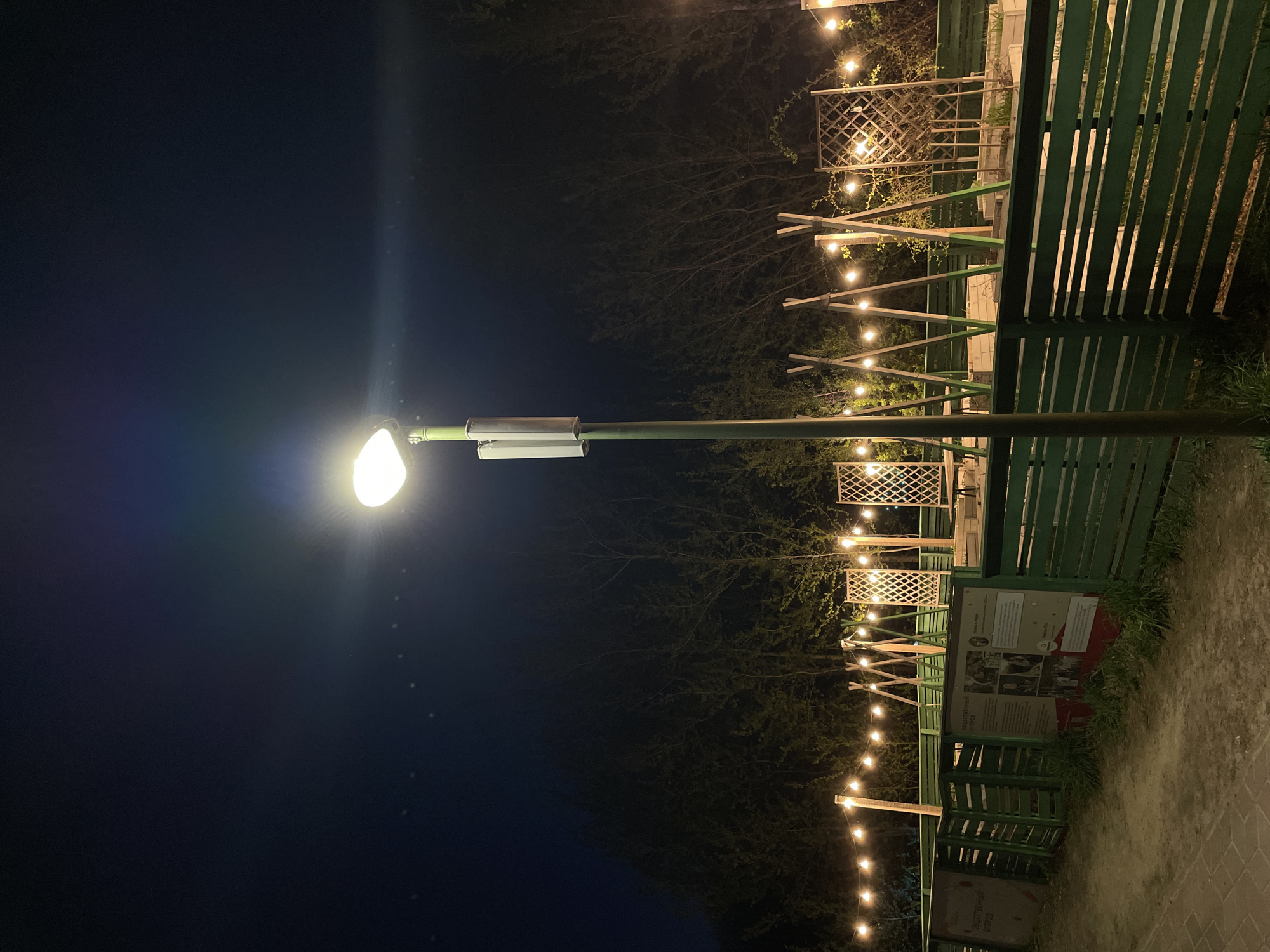 The image shows an outdoor scene at night in Kazan, Russia. A brightly lit street lamp dominates the left side of the frame. To the right of the lamp, a series of string lights stretches into the distance, suspended above a green wooden fence and trellis structures. A sign with text and images is visible along the fence. The background consists of trees and a dark sky. The ground appears to be a paved pathway.