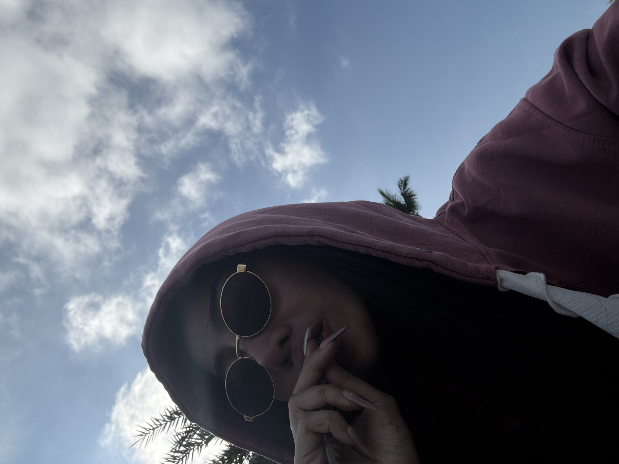 A low-angle shot shows a person wearing a burgundy hoodie with white drawstrings, round gold-rimmed sunglasses, and long artificial nails. The person is holding a thin object to their lips. The background includes a blue sky with scattered white clouds and the fronds of a palm tree. The location is implied to be outdoors in a sunny environment. The city is unknown.