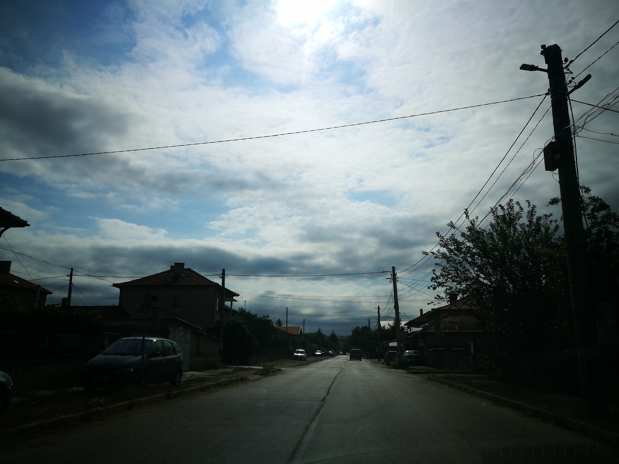 The image depicts a street scene in the city of Ihtiman, Bulgaria, under a cloudy sky with patches of sunlight breaking through. The perspective is from a vehicle traveling down the road. On either side of the asphalt road are houses with tiled roofs and some trees and vegetation. Utility poles with numerous wires are visible along the street. Several cars are parked or in motion on the road. A dark-colored hatchback is parked on the left side of the road in the foreground. The overall lighting suggests it is daytime, but the cloud cover creates varying light and shadow conditions.