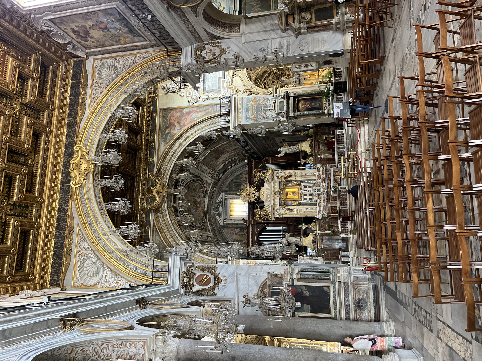The image shows the interior of a church in Rome, Italy. The ornate ceiling features gilded decorations, frescos, and multiple crystal chandeliers. Rows of wooden chairs line the floor. A person lies near a stone pillar. The ornate altar is centered in the background.
