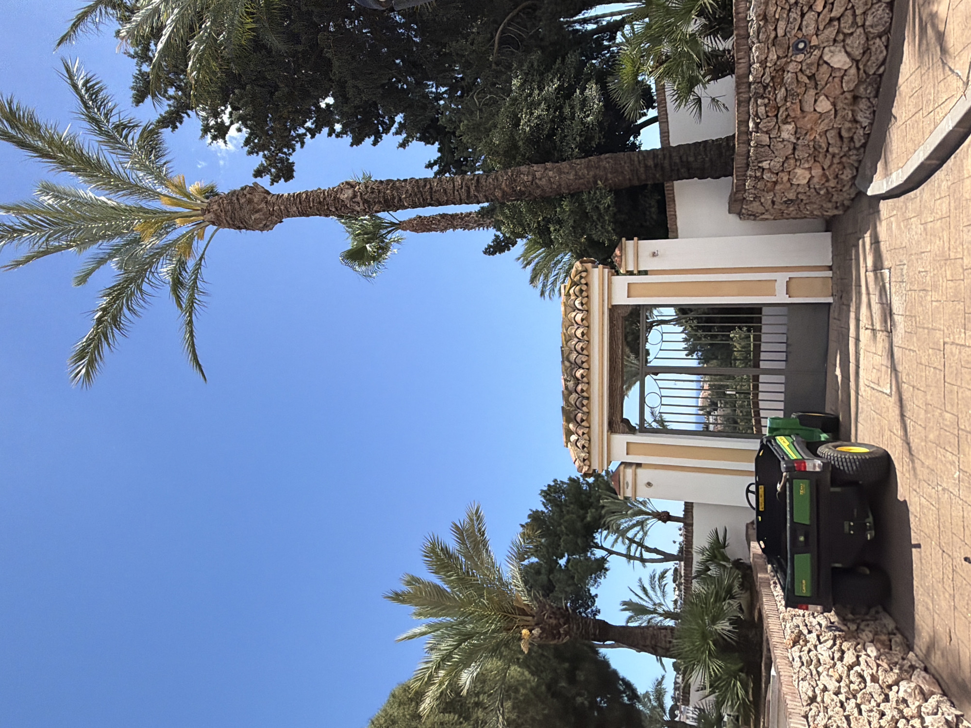 The image shows a scene in Benalmádena, Spain. A paved walkway leads to a gated entrance framed by white pillars with tan accents and a tiled roof. A green John Deere Gator utility vehicle is parked on the walkway, next to a low stone wall. Palm trees are present on both sides of the structure, and other trees and foliage can be seen in the background. The sky is clear and blue.