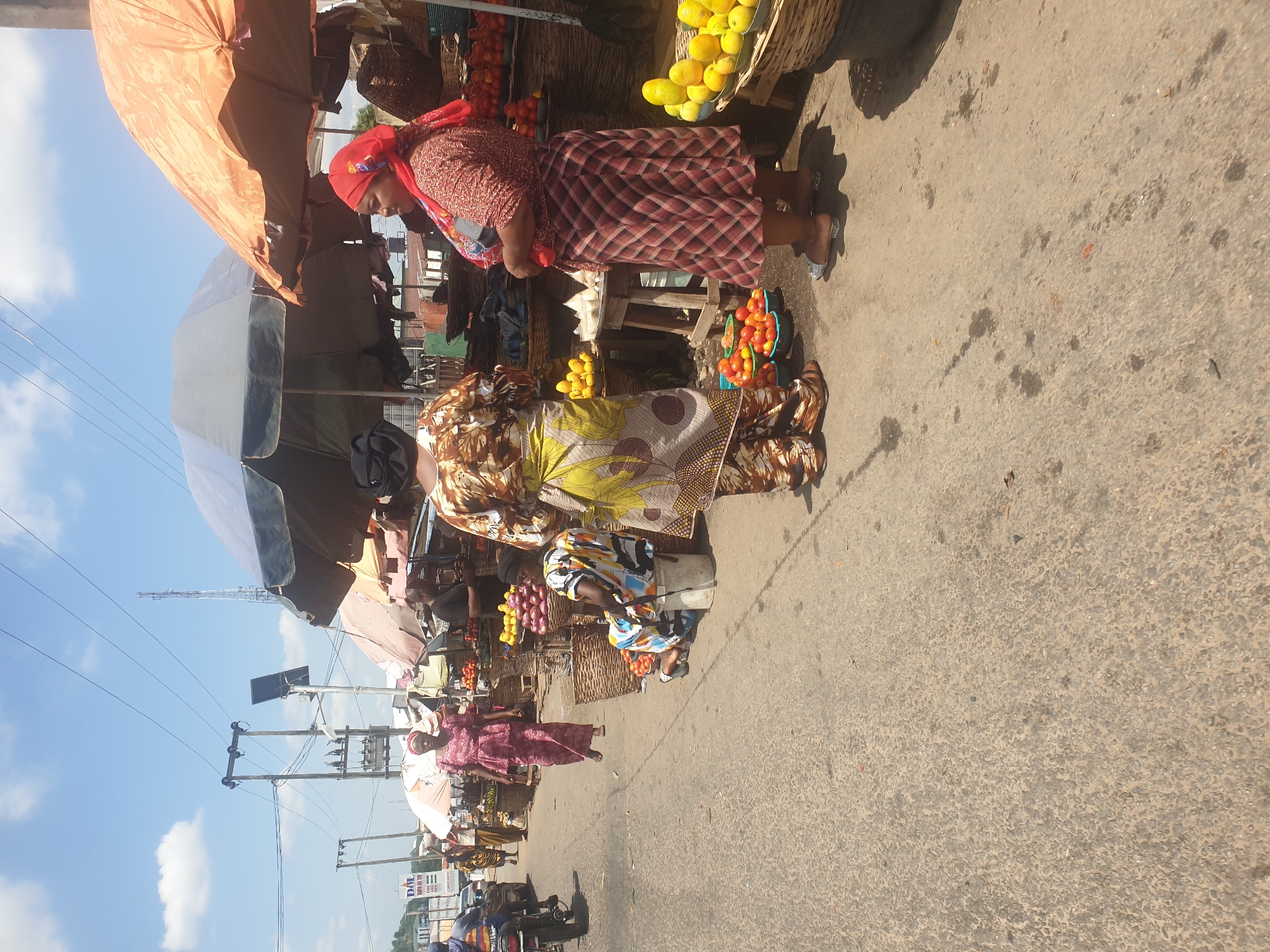 The image depicts a bustling marketplace in Lokoja, Nigeria. Several women stand at stalls displaying fruits and vegetables. One woman, wearing a red headscarf and a red and white plaid dress, appears to be a customer. Another woman, wearing a patterned dress with yellow and brown hues, stands behind a stall with baskets of produce. Other vendors and customers are seen further down the street, under umbrellas shielding them from the sun. The street is paved, and electrical poles with wires are visible against a bright blue sky with few clouds. Motorbikes are parked or moving near the edge of the market. The overall scene is typical of a busy market day in a Nigerian city.