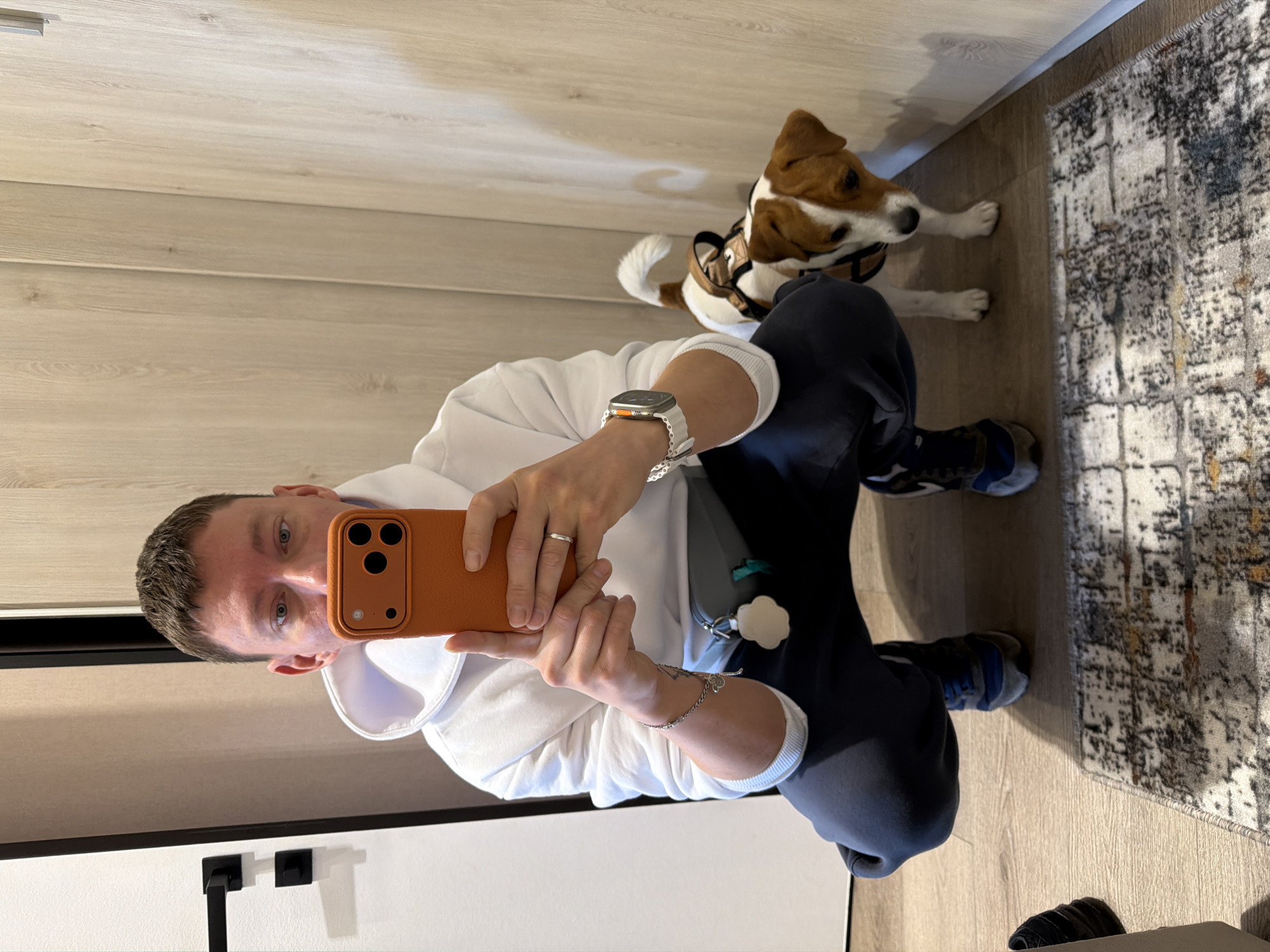 A Caucasian man, wearing a white hoodie and dark sweatpants, squats down for a selfie in what appears to be a residential entryway in Moscow, Russia. He holds an orange smartphone in his right hand, angled to capture his reflection in a light-colored wooden paneled wall to his right. A small Jack Russell Terrier wearing a brown harness stands to his right, looking towards the man. The man has short, brown hair and light blue eyes. He is wearing a ring on his left ring finger and a silver watch on his left wrist. A gray fanny pack is worn around his waist. To the left of the man, a white door with black hardware is visible. In the foreground, a grey and white patterned rug lies on a wooden floor.
