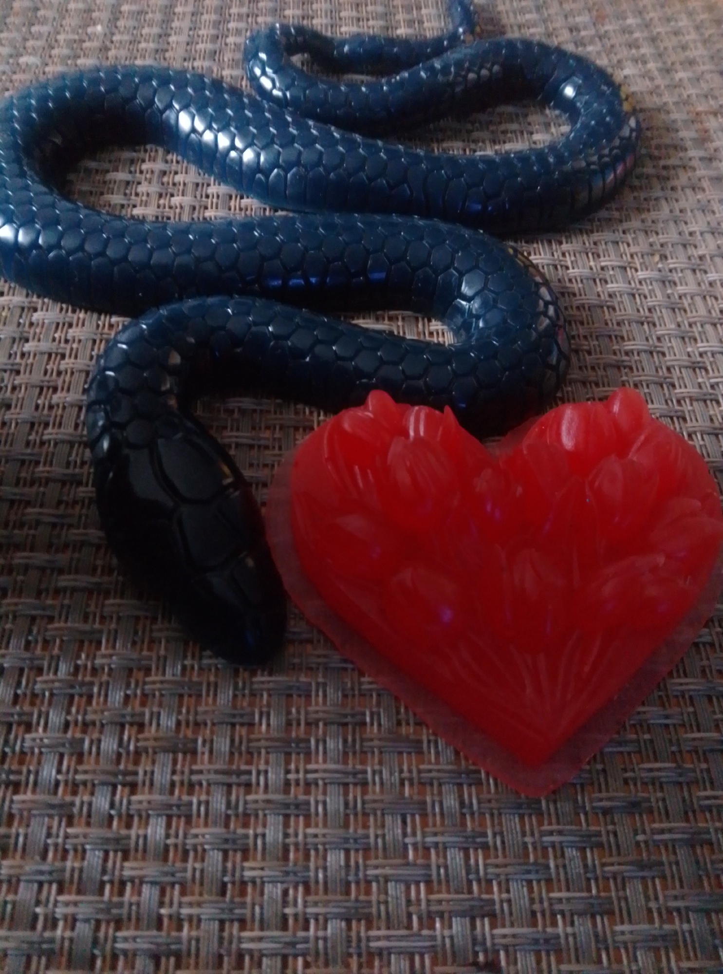 The image shows a coiled, dark blue, segmented snake toy placed next to a red, translucent heart-shaped object on a woven placemat. The snake's head is visible and appears black. The background is a textured, light brown and grey woven surface, likely a placemat. The lighting suggests an indoor environment. There are no people present in the image. The location in the city of Wedel, Germany is not discernible from the visual information provided.