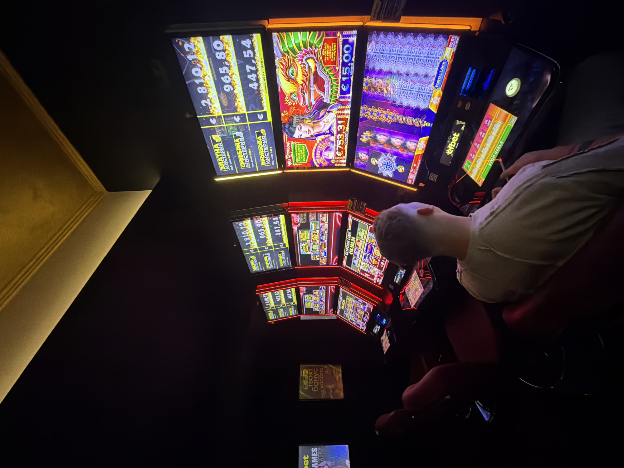 The image depicts the interior of a dimly lit gambling establishment, likely a casino or betting shop, in Veliko Tarnovo, Bulgaria. Several slot machines are prominently displayed, their screens illuminated with vibrant graphics and numbers. One machine on the right shows a prize of €753.31, with another displaying €15.00. Above these, a screen shows jackpot values of 2,810.12, 965.80, and 447.54. Another machine to the left displays the text "EGYPTIAN LINK" followed by a numerical value. Two individuals are visible in the foreground. One person, facing away from the camera, is seated in a red chair, looking towards the slot machines. Their clothing is partially visible. Another person is also seated, with their back to the camera, wearing a light-colored t-shirt, also facing the slot machines. The environment is dark, with a golden-colored ceiling feature visible in the upper portion of the image. A sign with Bulgarian text, reading "ТВОЯТ БОНУС НА КАСАТА" (Your Bonus at the Cashier), is mounted on the wall to the left, alongside a screen displaying the word "GAMES". The overall atmosphere suggests a typical setting for gambling activities.
