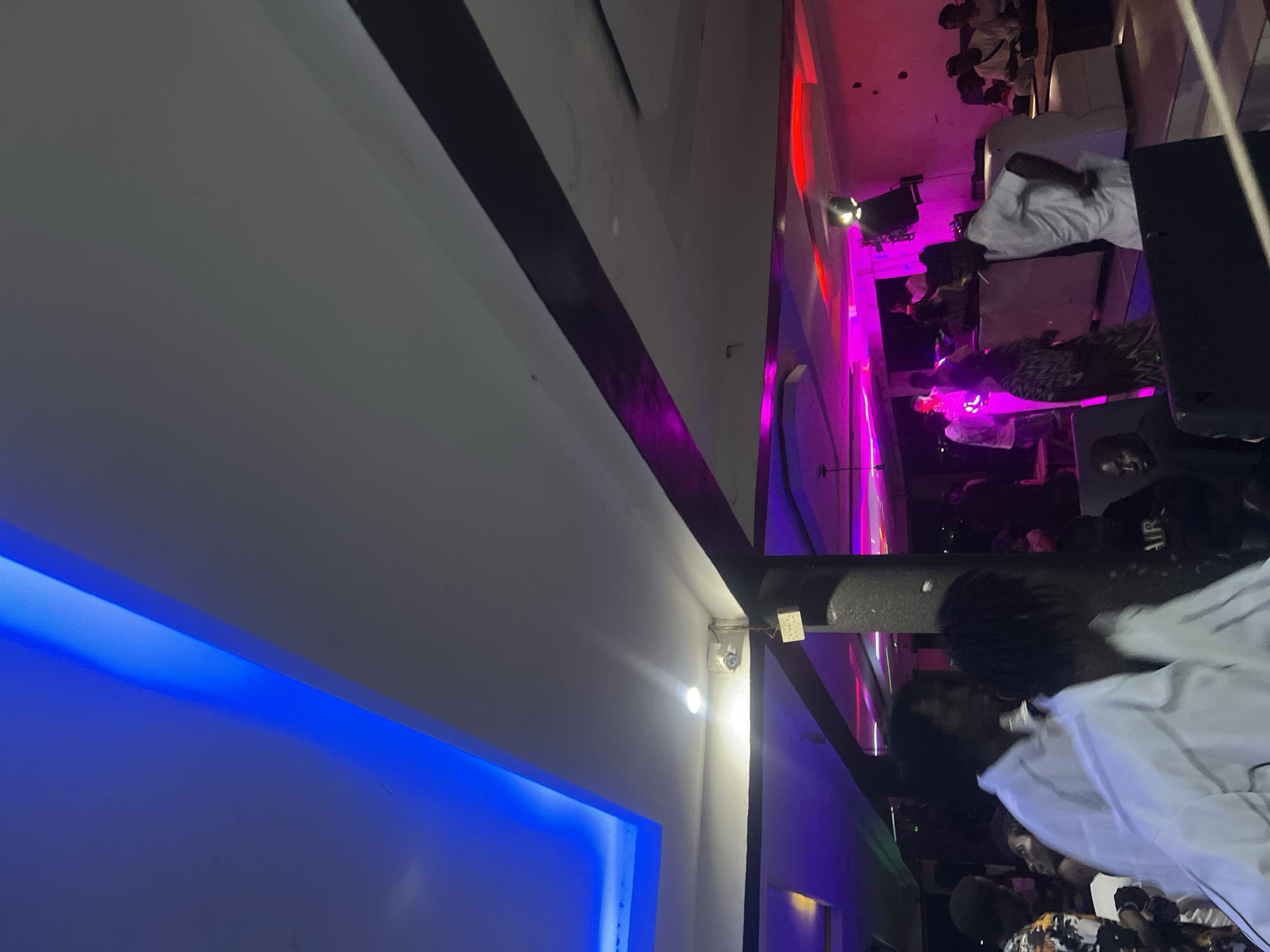 A slightly distorted perspective image taken inside a building, probably in Howayi, Nigeria, showing people at a gathering or event. The image is primarily oriented towards the ceiling and upper parts of the room, with lighting prominently visible.
On the left side of the frame, a portion of a wall is illuminated with a blue light strip. A dark, thick strip extends diagonally across the wall. There is a security camera attached to the ceiling.
The right side of the image captures a scene with people, possibly at a social event or gathering. There is a concentration of individuals, some wearing light-colored clothing. Multi-colored lighting, predominantly red and pink hues, casts a vibrant glow on the scene.