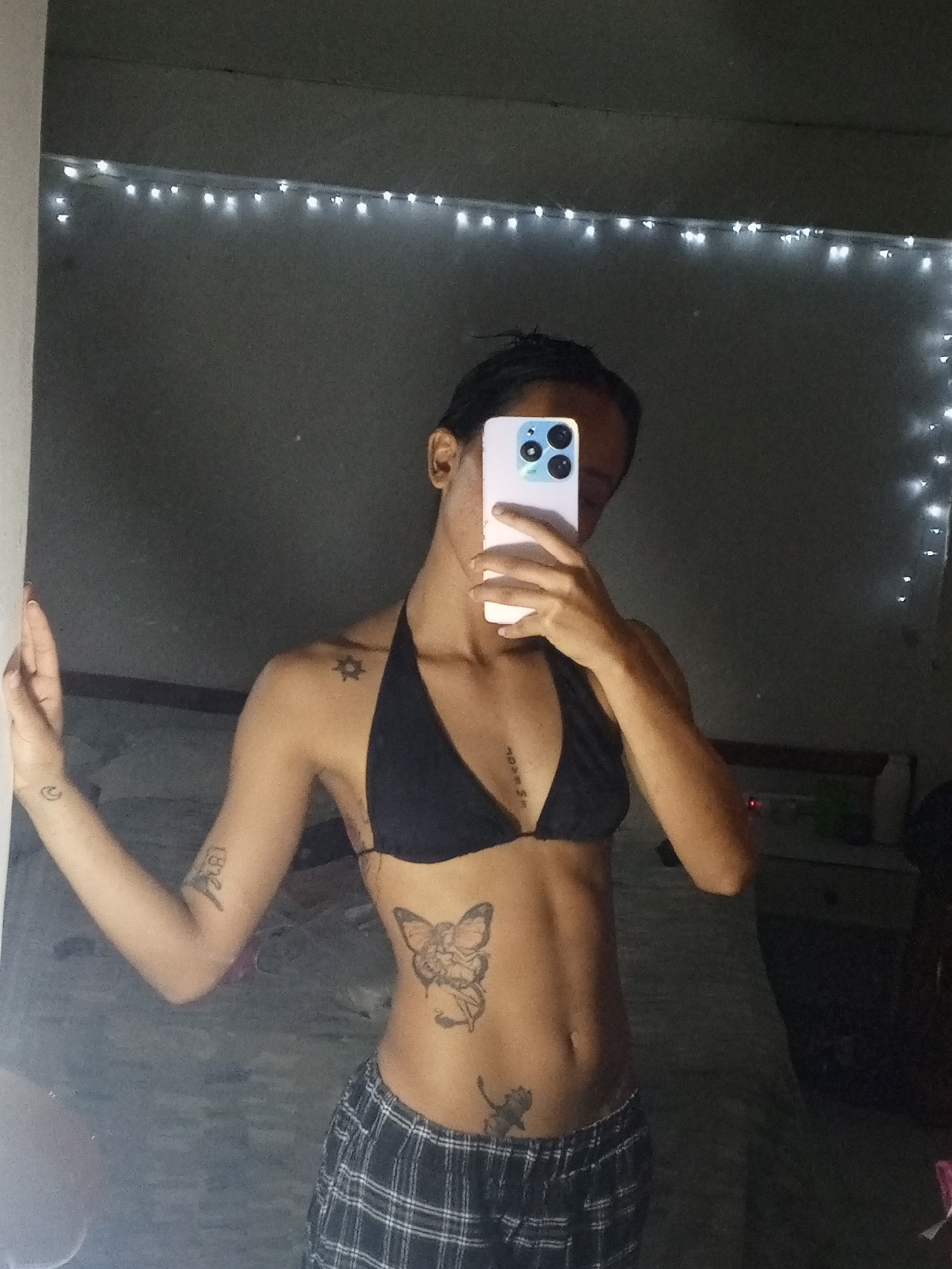 A person is taking a mirror selfie. The person is wearing a black bikini top and plaid pajama pants. They are holding a white smartphone in front of their face, obscuring their features. The person has multiple tattoos on their arms, torso, and stomach. The background shows a bedroom with a bed and string lights along the top of the wall. It is not possible to determine the location in Porlamar, Venezuela based on the available information.