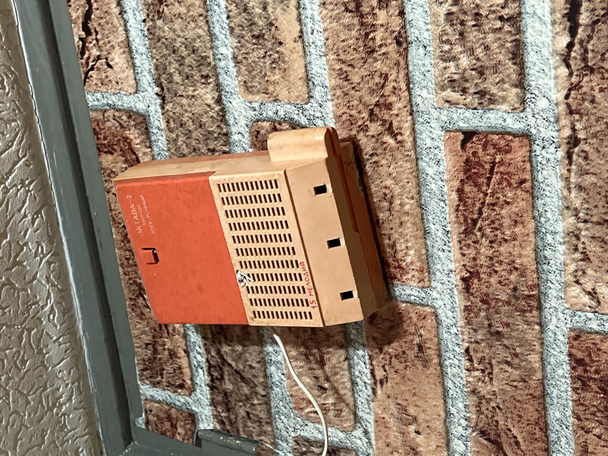 A vintage electronic device, likely an intercom or door chime unit, is mounted on a wall covered with brick-patterned wallpaper. The device, positioned slightly angled, is predominantly orange on its front panel and beige on its side and top sections. The front orange panel features a rectangular grill with numerous small perforations. A small, light-colored handle or latch-like element is visible on the upper part of the device.
Text in Cyrillic is visible on the device. On the orange front, it reads "ОКТАВА-2" (OKTAVA-2), with additional text below it that appears to be "3B.K. ГРОМКАЯ" (Zvuk Gromkaya), translating to "Loud Sound" or "Loudspeaker." Vertically aligned on the beige side panel, the text "15 МЕЛОДИЙ" (15 Melodiy) is clearly visible, indicating "15 Melodies." A thin white wire extends downwards from the bottom of the device.
The wall is adorned with wallpaper mimicking a brick pattern, featuring brown bricks with light gray mortar lines, suggesting a textured indoor surface. A dark gray vertical frame or structure is partially visible on the far left of the image. The scene is static, with no people or activity depicted, and the device's design suggests it is an older model, potentially from the Soviet era, fitting the Minsk, Belarus location context. The lighting indicates an indoor setting.
