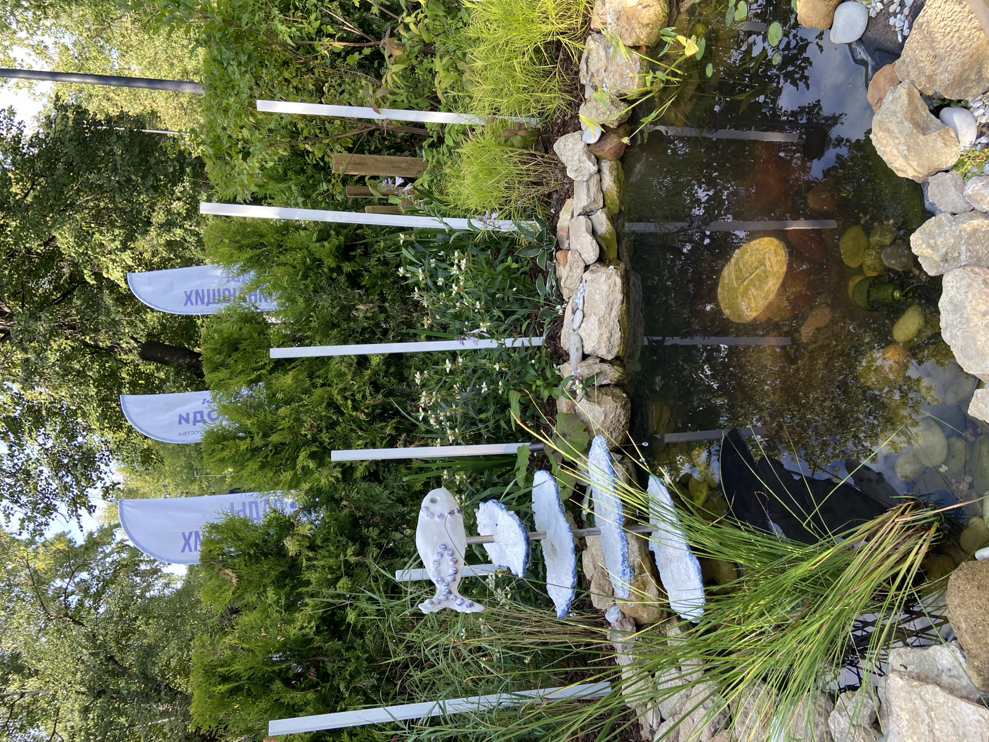 The image depicts an outdoor scene, likely a garden or park setting. In the foreground, a small pond is visible, surrounded by rocks and aquatic plants. A decorative element featuring a fish on a tiered stand is positioned near the pond. Behind the pond, there is a dense arrangement of green bushes and trees. Several white vertical poles are interspersed throughout the scene, and three large, white, curved flags with purple text are visible in the background, partially obscured by foliage. The overall impression is one of a landscaped outdoor space with decorative elements and advertising banners. The image does not contain any specific visual cues that would allow for its location to be identified as Moscow, Russia.