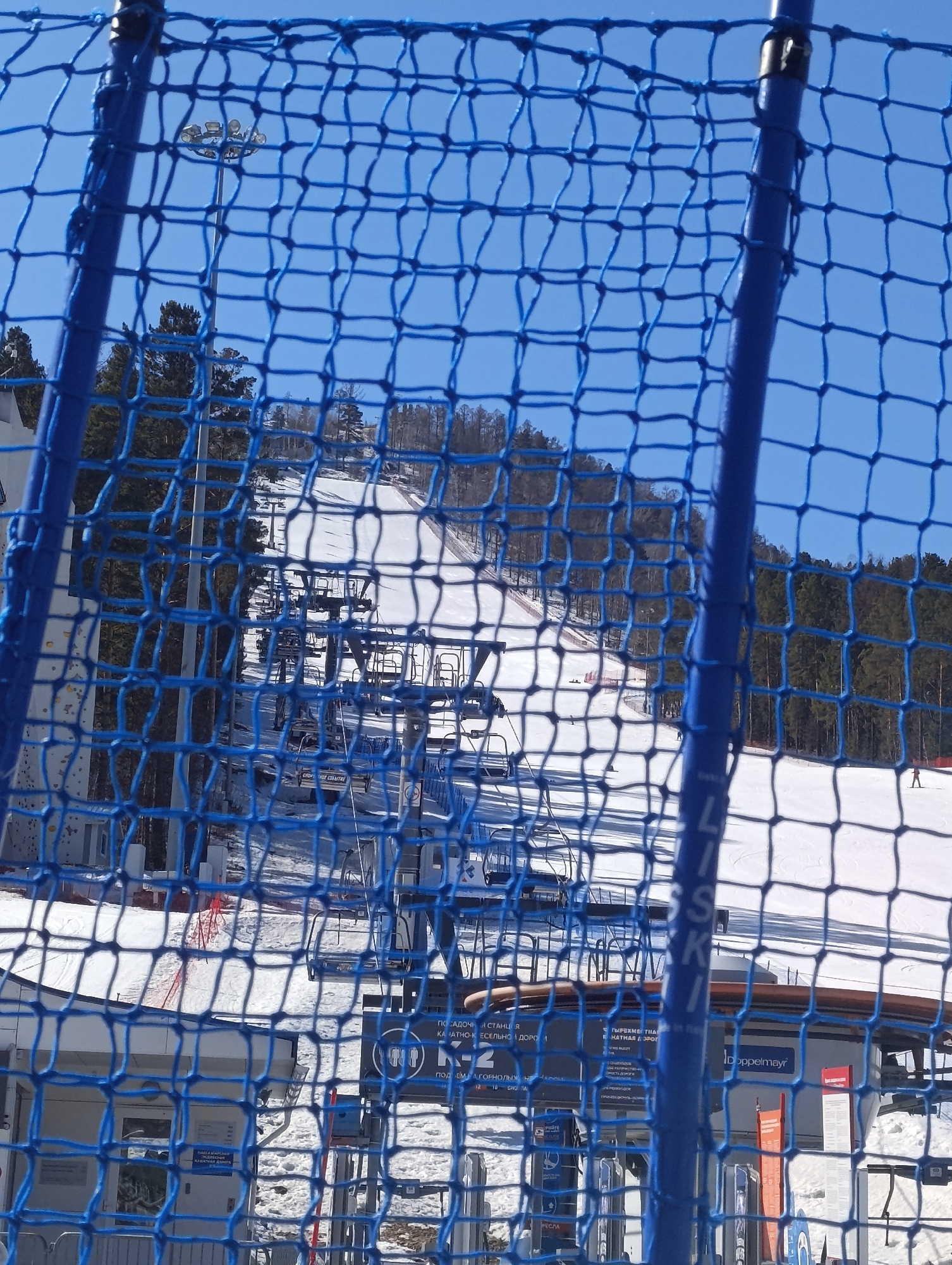Here is a matter-of-fact description of the image:
The image is a partial view through a blue safety net at the Bobrovy Log Funpark, a ski area in Krasnoyarsk, Russia. A snow-covered ski slope is visible beyond the net. A ski lift with several chairs runs up the slope. At the base of the ski lift, there are signs in Russian and the word "Doppelmayr" is visible. The sky is bright blue and mostly clear. The foreground shows the structure and mesh of the blue net. Trees line the edge of the ski run. In the distance, several people are skiing.
