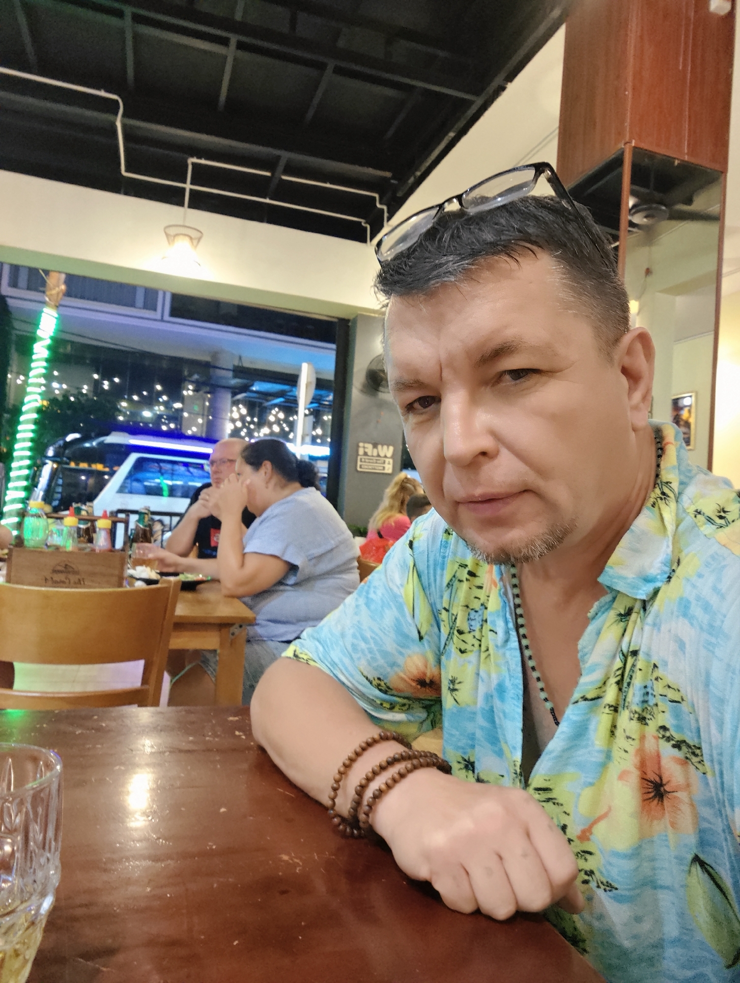 The image shows a close-up of a man sitting at a wooden table in an indoor restaurant setting. He is wearing a brightly colored, floral-patterned short-sleeved shirt and has glasses perched on top of his head. He is also wearing several beaded bracelets on his wrist. The man is looking directly at the camera with a neutral expression.
In the background, there are other people seated at tables, some of whom are partially visible. One man is wearing a black t-shirt and glasses, while a woman next to him is wearing a light blue t-shirt. The restaurant appears to be open to the outside, with greenery and lights visible through a large window. A bus with blue and white accents is parked outside. The lighting suggests it might be evening, as there are illuminated signs and lights from the street visible.
On the table in front of the man, there is a glass with a beverage and a wooden box with some bottles, possibly condiments. The overall ambiance seems casual, perhaps a meal or a drink at a restaurant in Nha Trang Ward, Vietnam. The image is a candid shot, capturing a moment during a meal or social gathering.