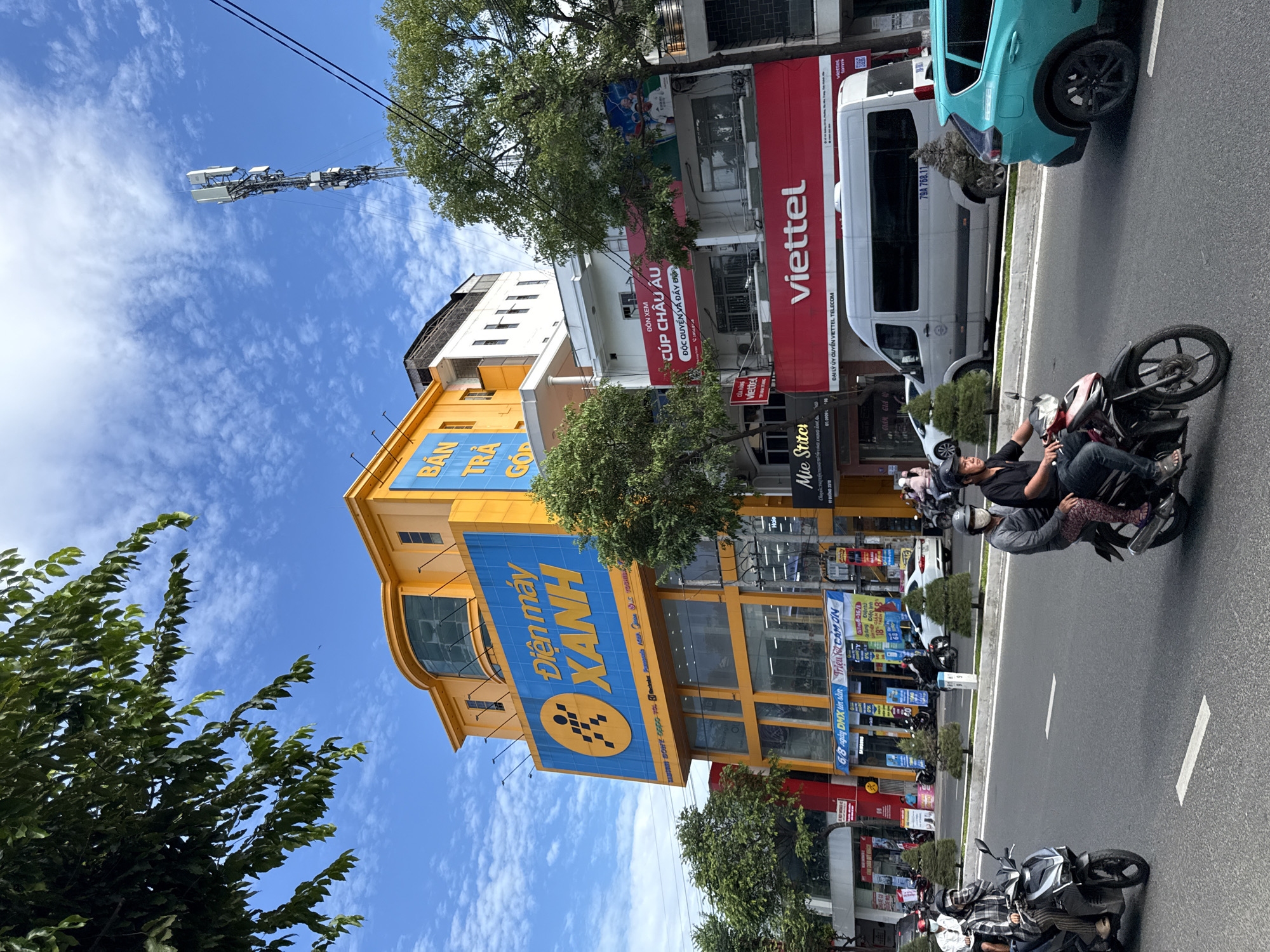 This is a street-level, eye-level shot taken in Nha Trang, Vietnam, on a partly cloudy day. The scene is dominated by a large, bright yellow building with a prominent blue sign that reads "Điện máy XANH" (Green Electronics), a popular electronics retailer in Vietnam. The building has multiple floors, large windows, and a curved glass facade on the upper level.
To the right of the yellow building are other commercial establishments, including one with a large red sign that says "viettel", indicating another prominent telecommunications company. Several trees line the sidewalk, providing shade and greenery.
The street is busy with traffic. Several motorbikes are visible, with riders wearing helmets and some with passengers. One motorbike in the foreground shows two people, both wearing face masks. A white van with the license plate "79A 768.11" is parked on the right side of the street. A teal-colored SUV is also visible on the right. The road has white lane markings. The overall impression is of a bustling urban environment on a bright day.