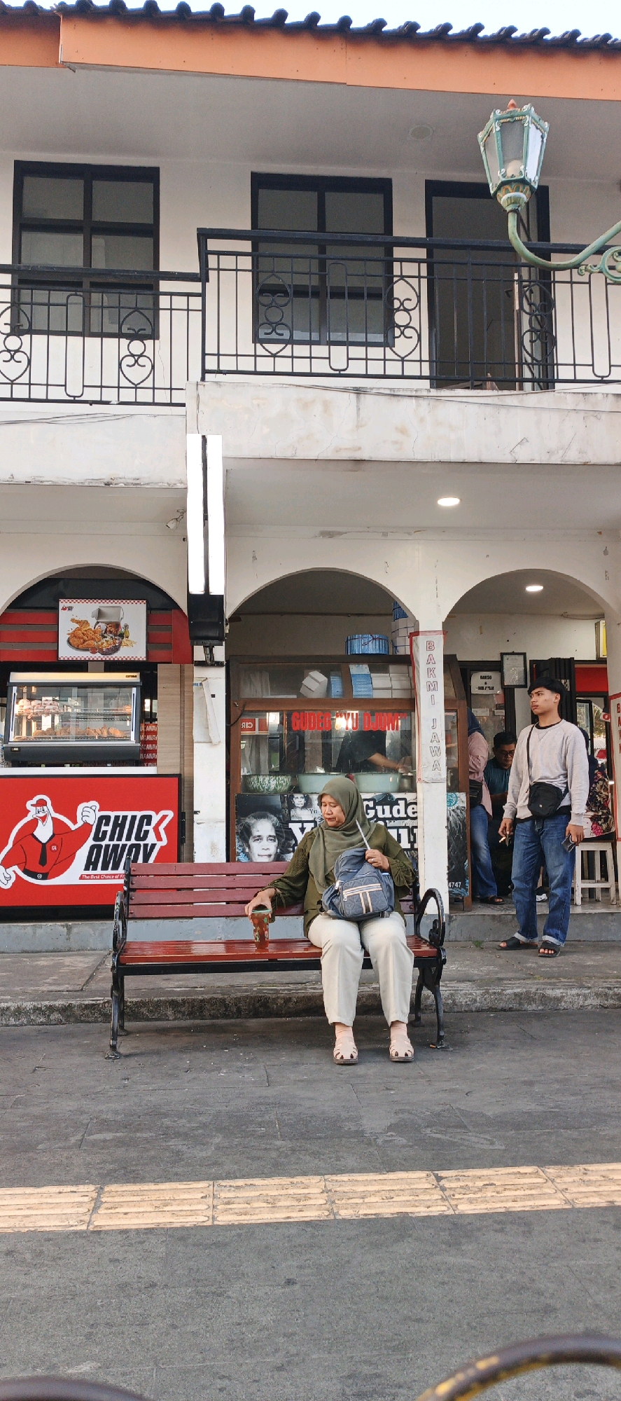 The image depicts an outdoor scene in front of a building with shops. A woman wearing a green hijab and olive green long-sleeved shirt is seated on a red wooden bench. She has light-colored pants and sandals on and is holding a brown cup in her right hand. A grey backpack is on her lap. To her right, a young man in a grey sweatshirt and blue jeans stands looking to the right. He has a black bag slung across his chest. Behind them, the building has arched entrances to shops. One shop has a display of fried chicken with a sign that reads "CHIC AWAY". Another shop has a sign with Indonesian text that appears to be "GUDEG YU DJO..." and "BAKMI JAWA". The area appears to be a street or pedestrian walkway.