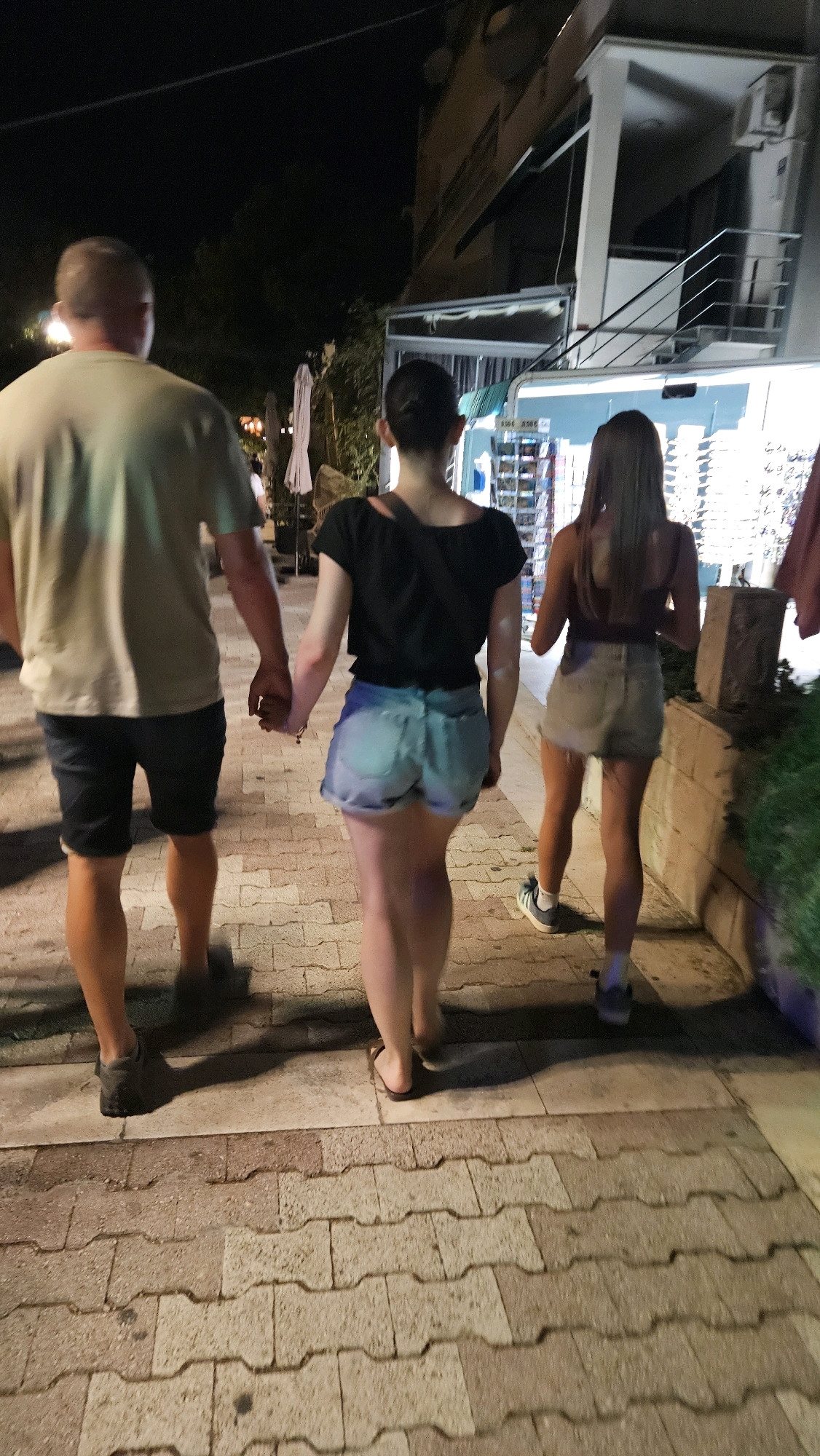 A man and two women are walking away from the camera on a paved sidewalk at night. The man, on the left, is wearing a light-colored t-shirt and dark shorts, holding hands with the woman in the middle. The woman in the middle is wearing a black crop top and light blue denim shorts, with her hair pulled back in a bun. The woman on the right is wearing a brown tank top and light-colored denim shorts, with her hair down. They are walking past a building with a lit storefront and an outdoor display. The sidewalk is made of interlocking concrete pavers. The background is dark with some trees and building lights visible. The location is identified as Općina Tučepi, Croatia.