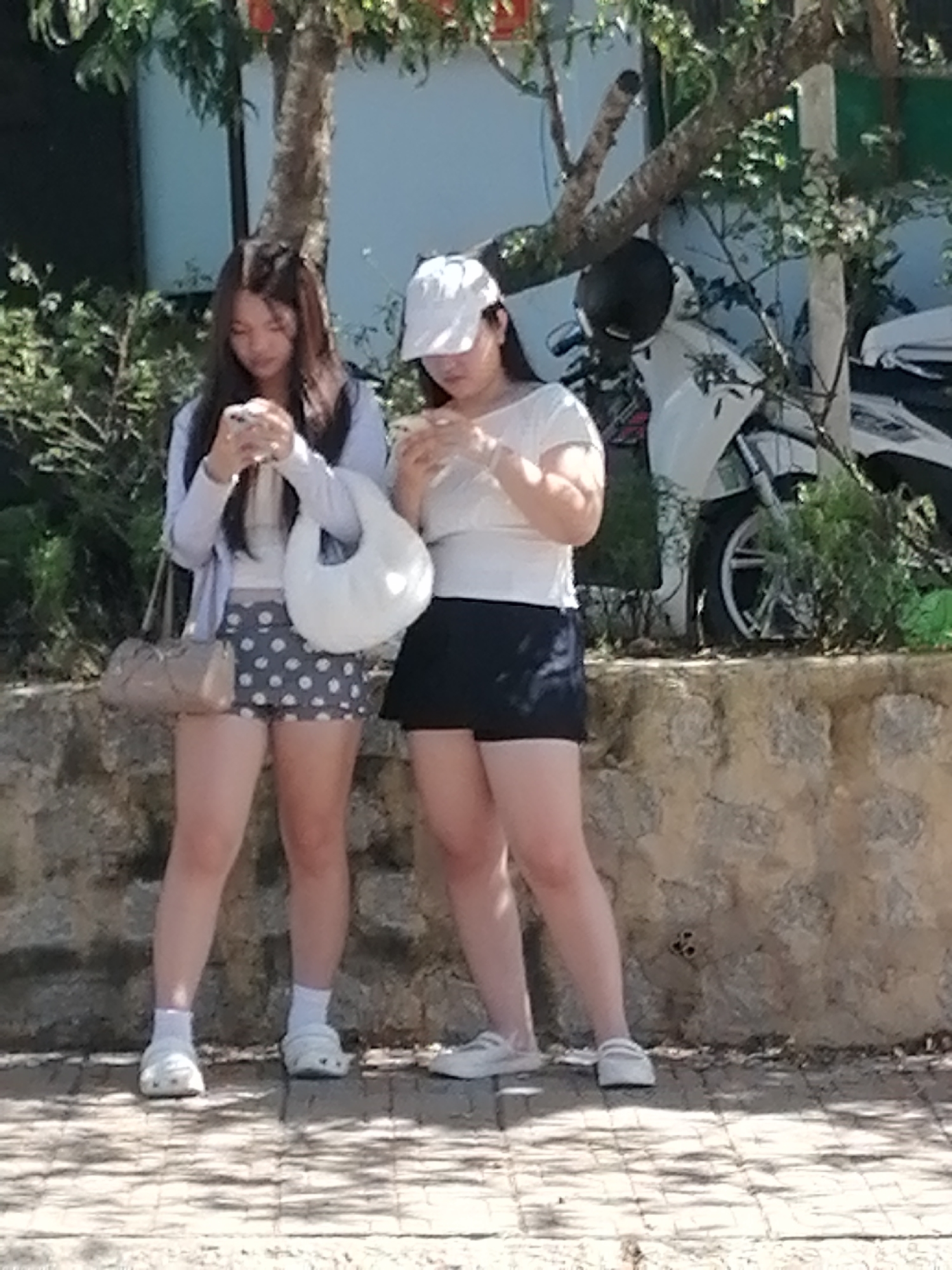 The image depicts two young women standing outdoors, possibly in Xuân Thành, Vietnam, based on the context. The woman on the left has long, dark hair and is wearing a light blue cardigan over a white top, a polka-dotted skirt, white socks, and white slip-on shoes. She is holding a beige shoulder bag and a white purse, and is looking down at her phone. The woman on the right is wearing a white t-shirt, a black skirt, and white slip-on shoes. She has a white baseball cap on and is also looking down at her phone. Behind them, a white scooter is parked. The background features a stone wall and lush green foliage.