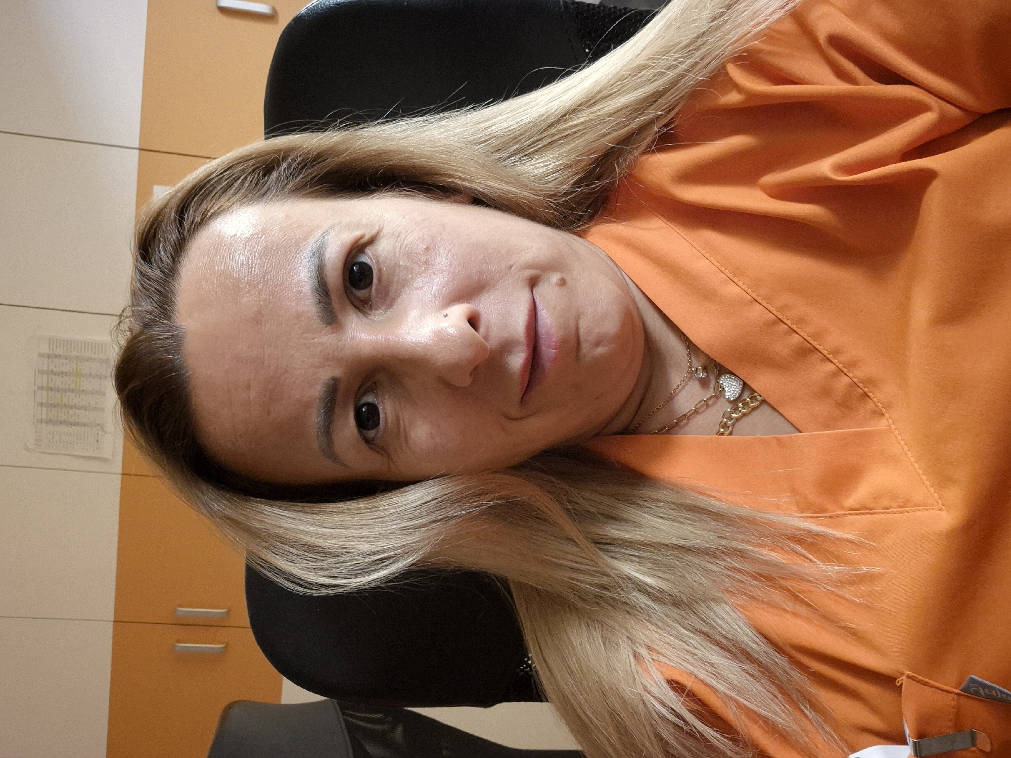 The image is a head and shoulders selfie of a blonde woman wearing an orange scrub top. She is sitting in a black chair, with orange cabinets and a white wall behind her. There is a piece of paper with a grid pattern on the wall. The woman has long, blonde hair and is wearing two necklaces, one with a diamond pendant and the other with a heart-shaped pendant. The lighting is bright and appears to be from an overhead source. The background is blurred, suggesting a shallow depth of field. The image quality is sharp, and the colors are vibrant. The woman's expression is neutral, with a slight smile.