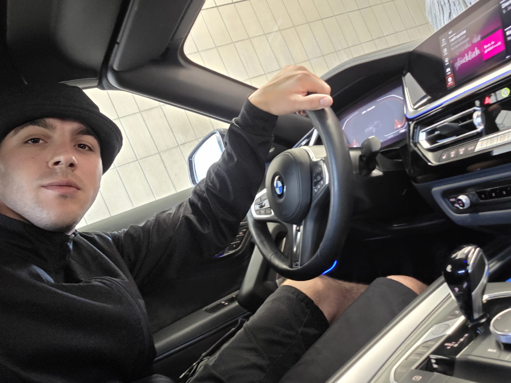 A young man wearing a black bucket hat and black long-sleeved shirt sits in the driver's seat of a car, holding the steering wheel. He is looking towards the camera, and his face is partially visible. The car's interior is modern, with a large digital dashboard and infotainment screen displaying music information. The steering wheel has a BMW logo in the center. The image appears to have been taken indoors, possibly in a dealership or showroom, as indicated by the tiled floor and overhead lighting. The man is wearing black shorts. The transmission shifter is visible in the foreground. The car's ambient lighting is blue.