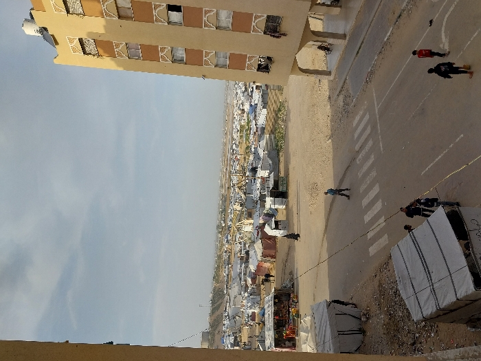 An elevated view captures an expansive, informal settlement in Khan Yunis, Palestinian Territory. The scene features a mix of temporary shelters and permanent structures under a light blue, hazy sky with scattered clouds.
In the immediate foreground, to the left, a partial view of a light-colored building's wall is visible, indicative of the vantage point. Below this, a large, rectangular, light-colored tent or covered structure is situated on dirt ground adjacent to an asphalt road. Another similar, slightly darker tent is partially visible further down.
To the right, a multi-story, tan-colored building rises, featuring multiple floors with rectangular windows framed by decorative, light-colored arched motifs. A white water tank and a solar water heater are visible on its roof. The ground floor of this building features three distinct arched open spaces, beneath one of which a person in a wheelchair is partially visible.
An asphalt road runs across the mid-foreground, curving slightly to the right, with a marked white-striped pedestrian crosswalk in the center. A dark cable or rope extends across the road.
Several individuals are present:
* A male figure in blue trousers and a light blue shirt walks left-to-right across the crosswalk.
* Three individuals are gathered near the tent structure on the left, one wearing a dark jacket and another with a backpack.
* Further right on the road, two younger male figures walk away from the camera; one in a red shirt and dark trousers, the other in a black hoodie and dark trousers.
* In the midground, near a makeshift stall, a person in a long dark garment walks away.
The midground is dominated by an extensive refugee camp comprised of countless tents and temporary shelters constructed from various materials, including white, grey, and beige tarpaulins, some with corrugated metal or wooden components. The ground throughout the camp is arid and sandy. A colorful market stall, displaying various goods under a red awning, is visible on the left side of the camp.
The background reveals more temporary structures extending to a low, light-colored hilly or mounded terrain, with additional distant structures dotting the horizon under the hazy sky.
