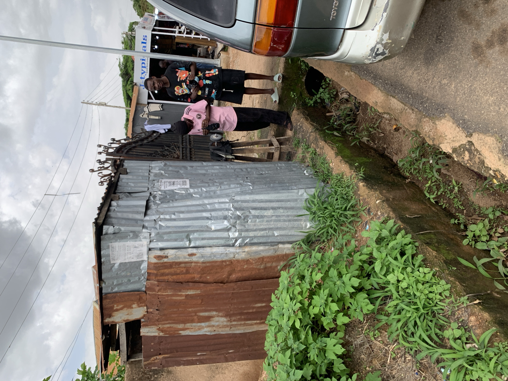 The image depicts two young men standing in an outdoor setting. One man is wearing a black t-shirt with a colorful graphic and black shorts, while the other is in a pink t-shirt and black pants. They appear to be in a somewhat dilapidated area, with a corrugated metal structure and a rusty gate visible behind them. A car is parked to their right, and a drainage ditch filled with green vegetation runs alongside the paved area. The sky is overcast, suggesting a cloudy day.
In the background, there's a glimpse of a business with signage that reads "Jtypi als". The overall impression is one of a casual encounter in an urban or semi-urban environment in Jos, Nigeria. The men's postures suggest they might be engaged in conversation or observing something. The presence of the car and the businesses hints at a lively, everyday scene, despite the slightly worn aesthetic of the surroundings. There are no strong indicators of a specific event, but the scene conveys a sense of normalcy.