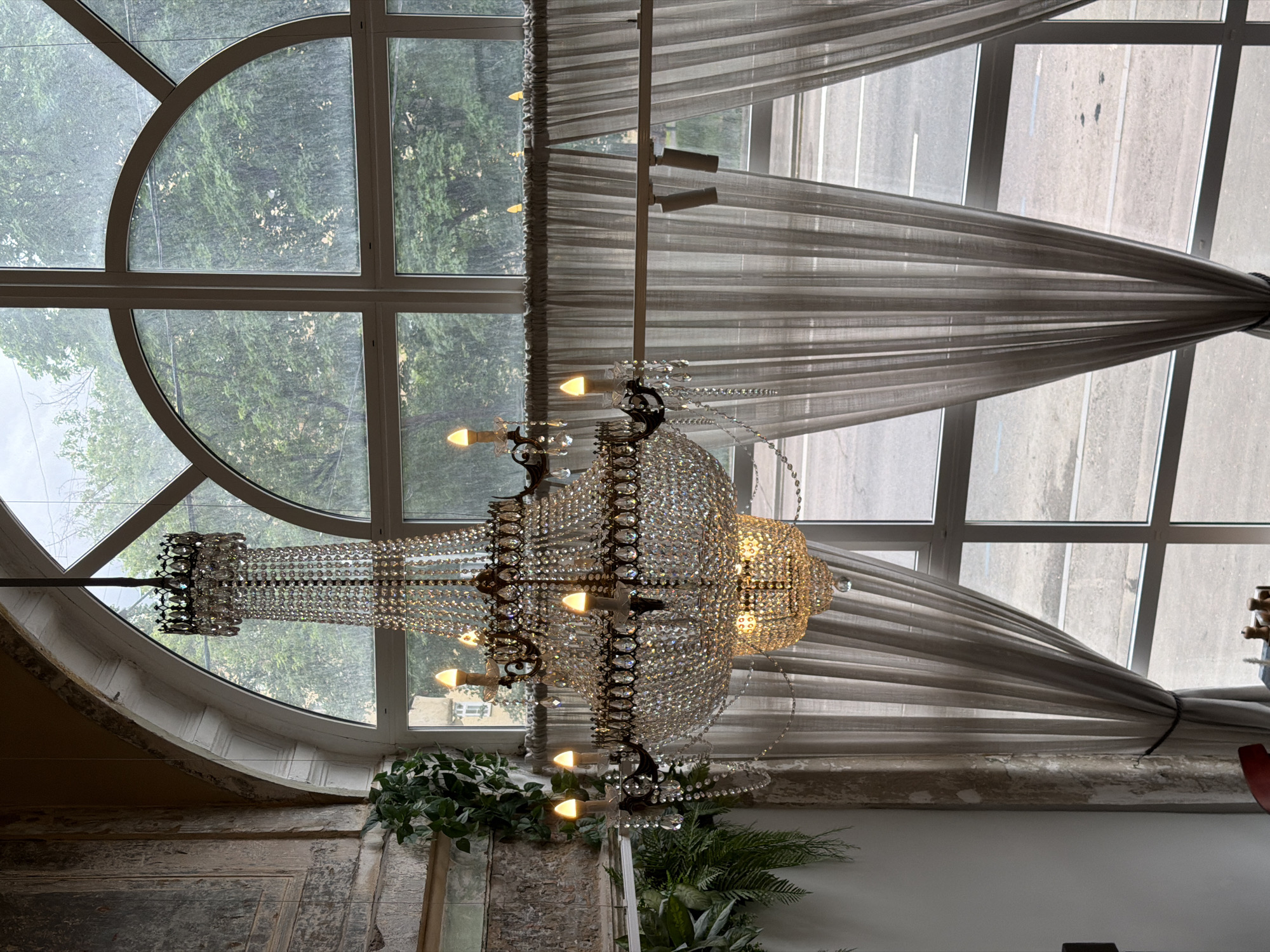 The image captures an interior scene in Moscow, Russia, notable for a large, ornate crystal chandelier centrally positioned in front of a substantial arched window. The window is divided into numerous panes, with the upper portion forming a semi-circular arch. The view through the window shows dense green foliage, suggesting trees or a park-like environment, contrasted by a road visible through the right side of the window. Grayish sheer curtains are draped on either side, creating a symmetrical frame.
The chandelier itself features multiple tiers of crystals and illuminated bulbs. The room's interior hints at aged architecture with weathered-looking walls. Small indoor plants add a touch of greenery to the space.