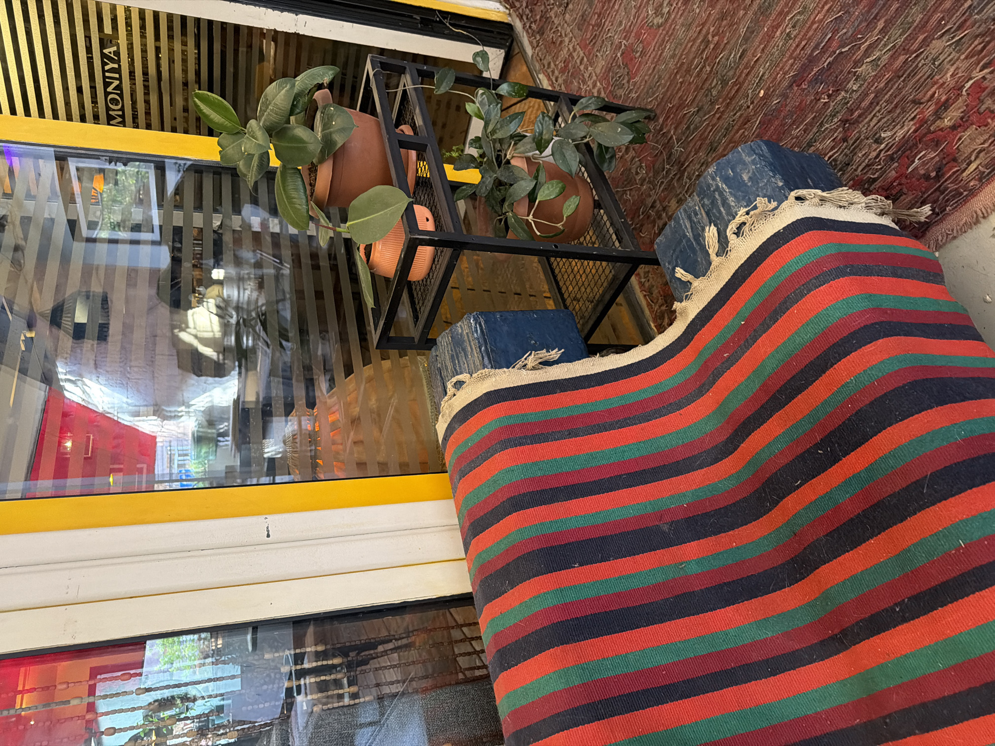 The image captures an outdoor scene in Yerevan, Armenia. A section of a storefront is visible, featuring a window with a yellow frame and the word "MONIYA" displayed vertically. Two potted plants sit on a black metal shelf in front of the window. The pots are terracotta-colored. Below the shelf, two blue blocks support the shelf. A striped rug with green, red, and black stripes is draped nearby. Another rug in varying shades of brown is partially visible in the background.