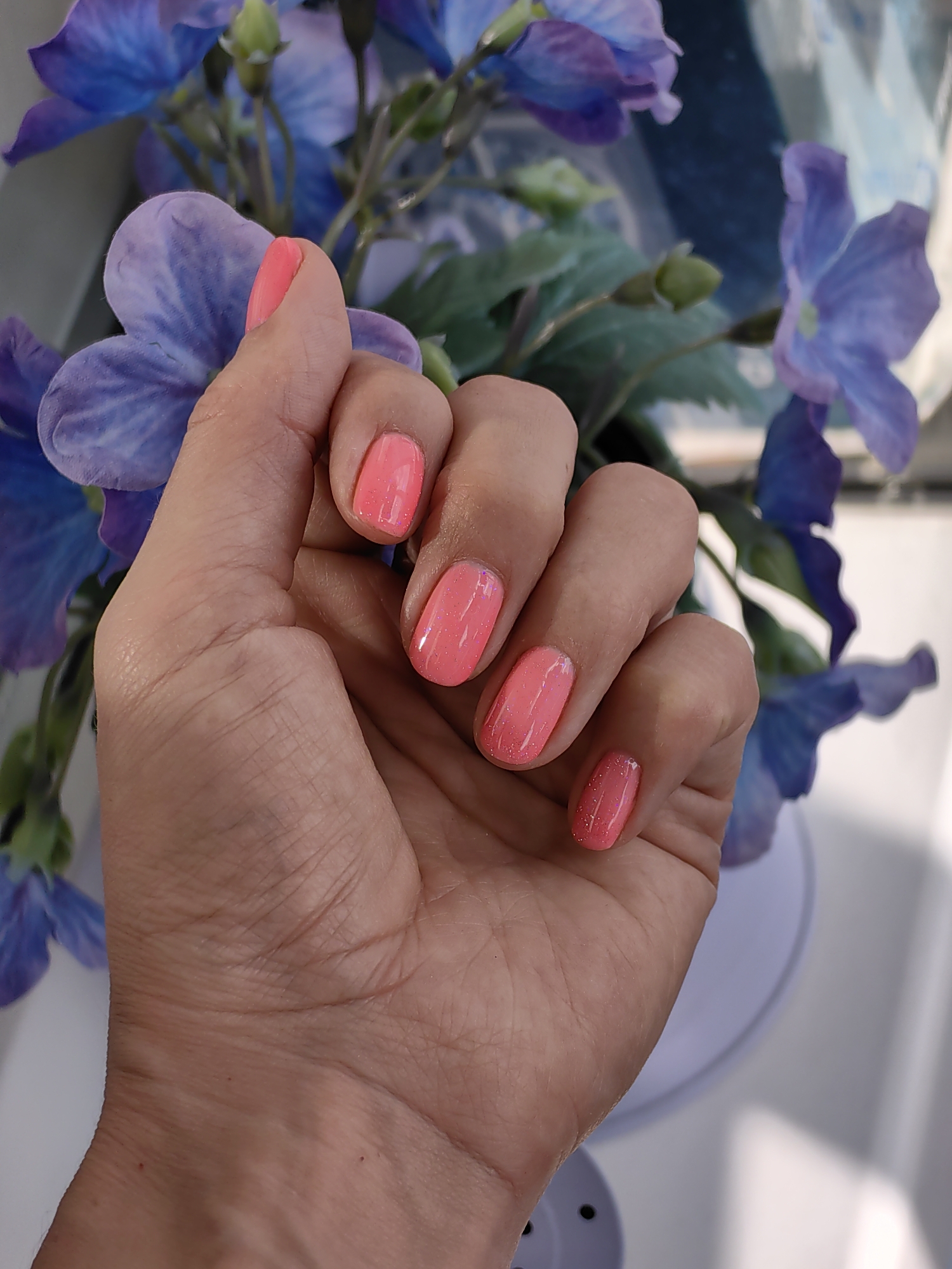 The image shows a close-up of a person's left hand with neon pink nail polish, in front of a blurry background of blue and purple flowers. The hand is resting on a pale purple object with a textured surface and black holes. The scene is indoors, with natural light coming from the right. There are no identifiable landmarks or elements that would suggest a location in Fanipal, Belarus.