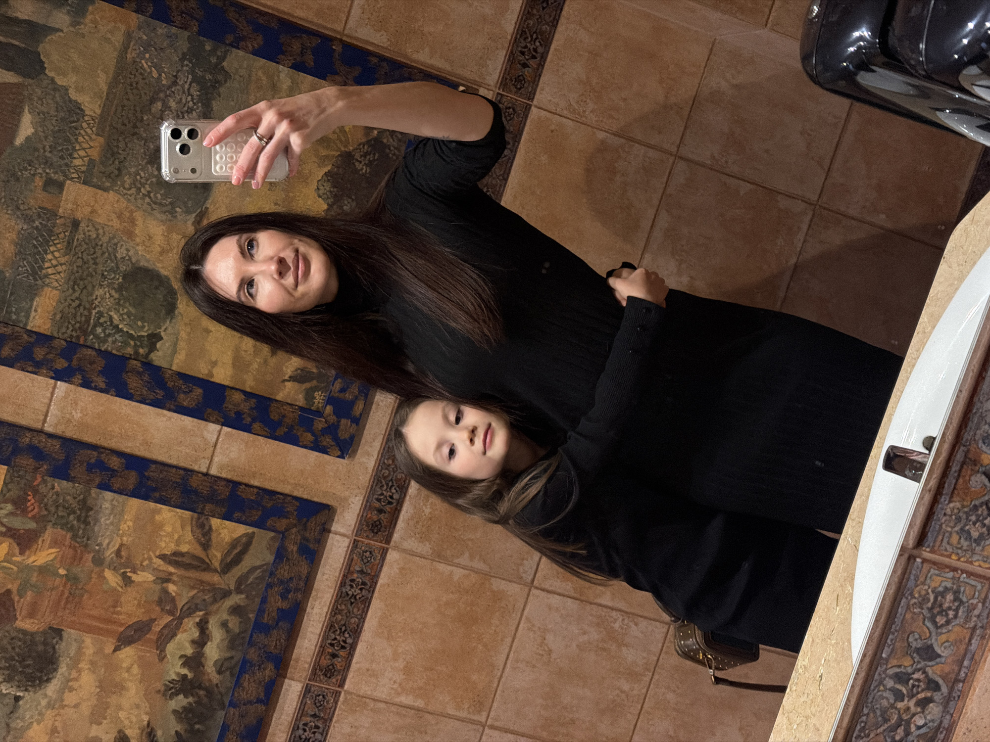 A woman and child are taking a selfie in a bathroom with brown tile floors and walls in what appears to be Perm, Russia. The woman is holding a smartphone in her left hand while embracing the child in a black dress. The floor and walls have decorative tile with floral and geometric patterns. The bathroom appears to have a sink with a white basin. The image appears to be taken near a public bathroom, probably in some tourist attraction in the city of Perm.