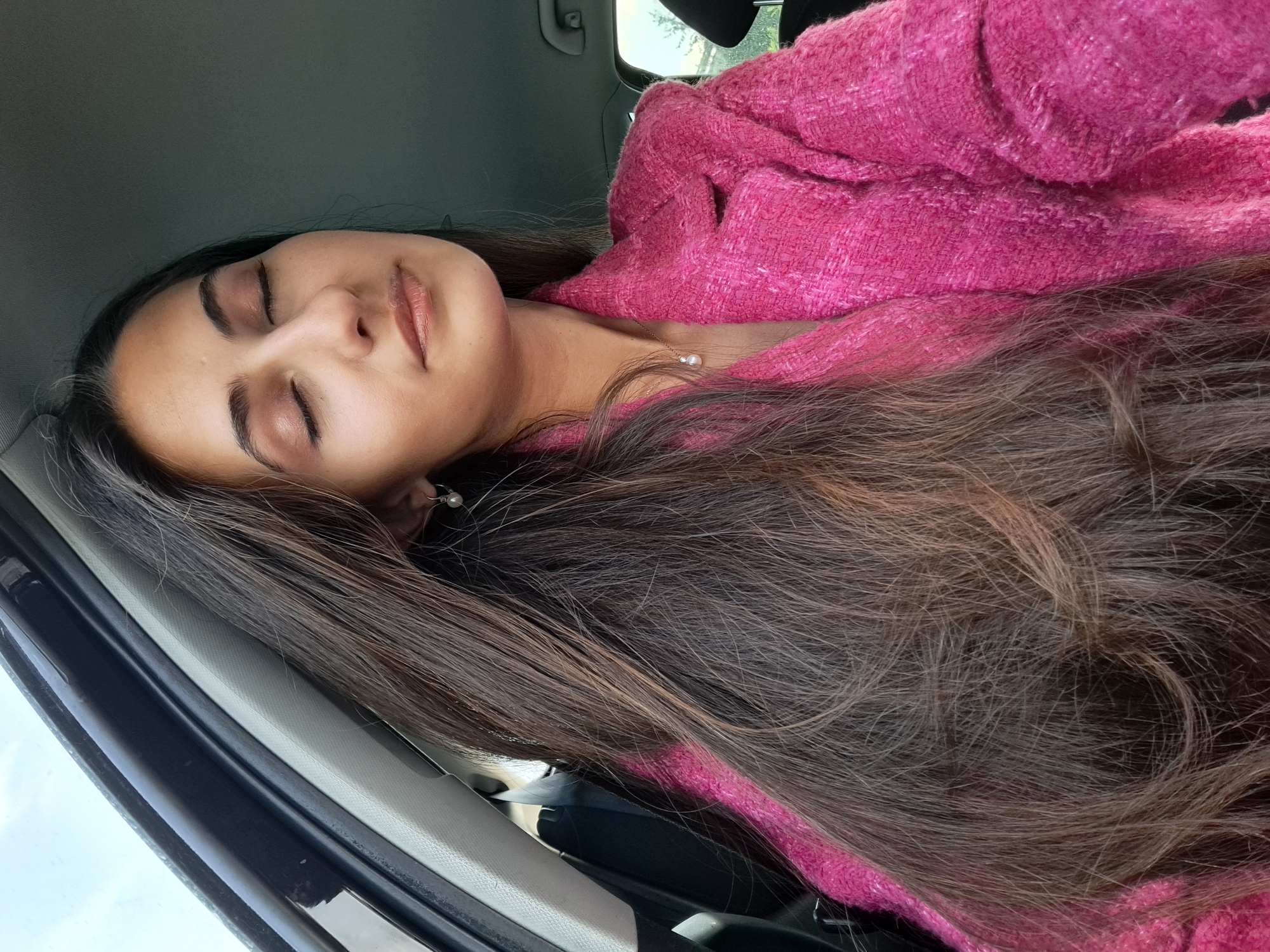 The image captures a young woman resting or sleeping inside a car, viewed from an overhead perspective. Her head is tilted back, with her eyes gently closed, conveying a sense of peace or relaxation. Her long, dark brown hair spreads out around her head and shoulders. She is wearing a bright pink, textured jacket or top. A delicate gold chain necklace with a single pearl pendant is visible on her neck, and a small pearl earring can be seen near her ear.
The setting is the interior of a car, with parts of the car's structure, including a gray headliner and a dark door frame, visible around her. The lighting indicates it is daytime, with soft, diffused light illuminating her face from what appears to be a car window. There is no visible text in the image.