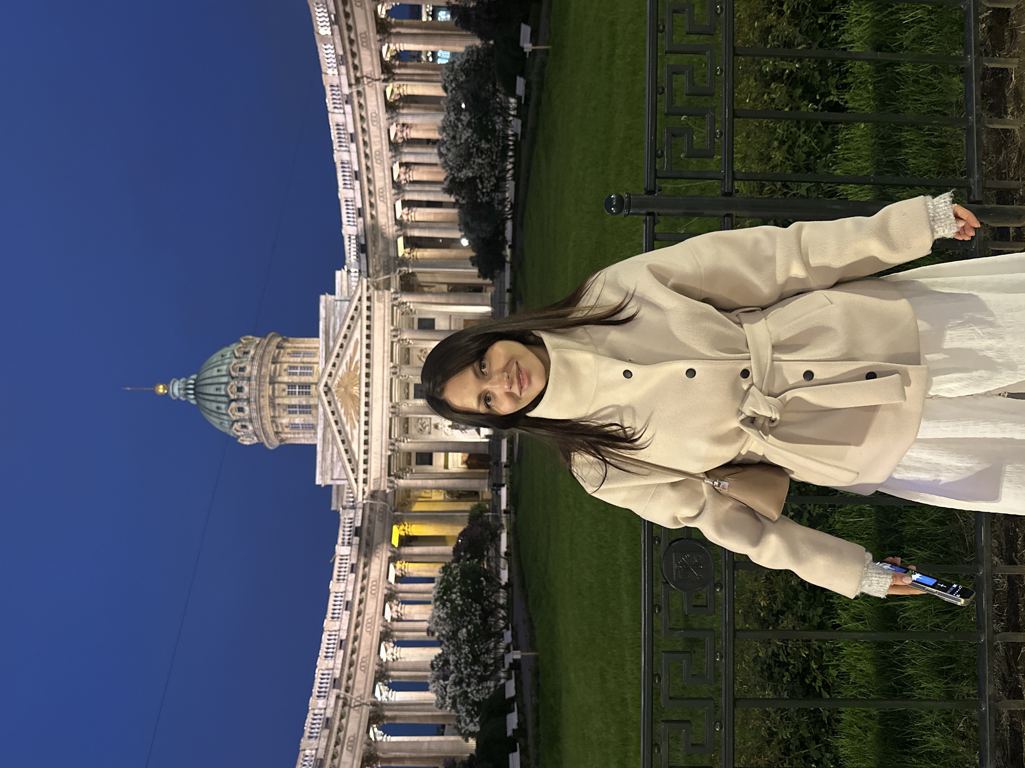 A medium shot shows a woman posing for a picture in front of the Kazan Cathedral in Saint Petersburg, Russia. She wears a beige belted coat, a white dress, and holds a smartphone in her left hand. The cathedral's colonnade and dome are illuminated against the night sky. A dark metal fence separates the woman from the grassy area in front of the cathedral.
